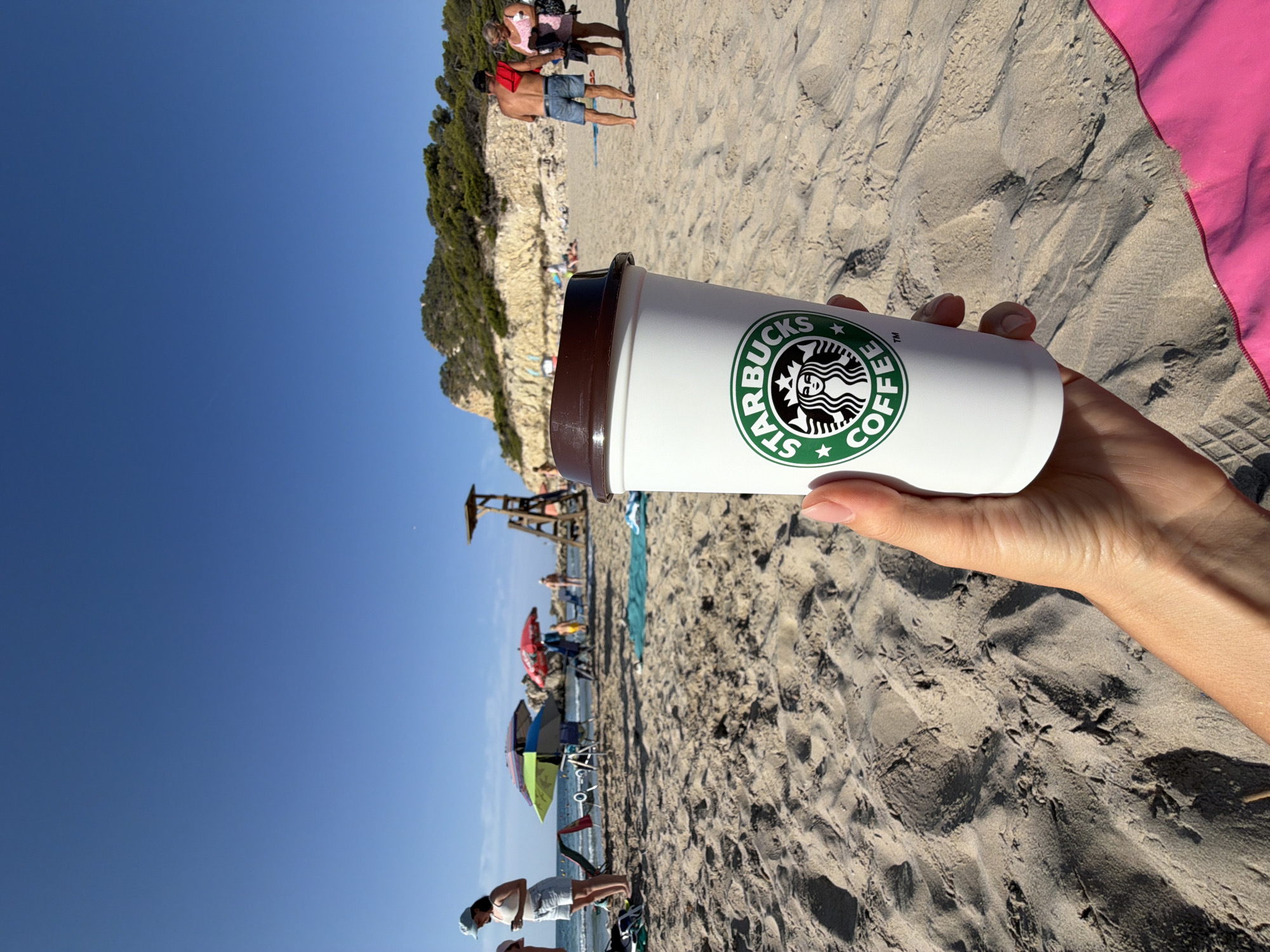 A first-person perspective shot shows a hand holding a white Starbucks coffee cup with its logo visible, set against a sandy beach with the ocean and a rocky, tree-covered hillside in the background. The sky is clear blue. In the mid-ground, several beach umbrellas, chairs, and people are scattered on the sand. A lifeguard tower is visible on the right side of the frame. The overall scene depicts a sunny day at the beach in Urbanització Cales I Talaies, Spain.