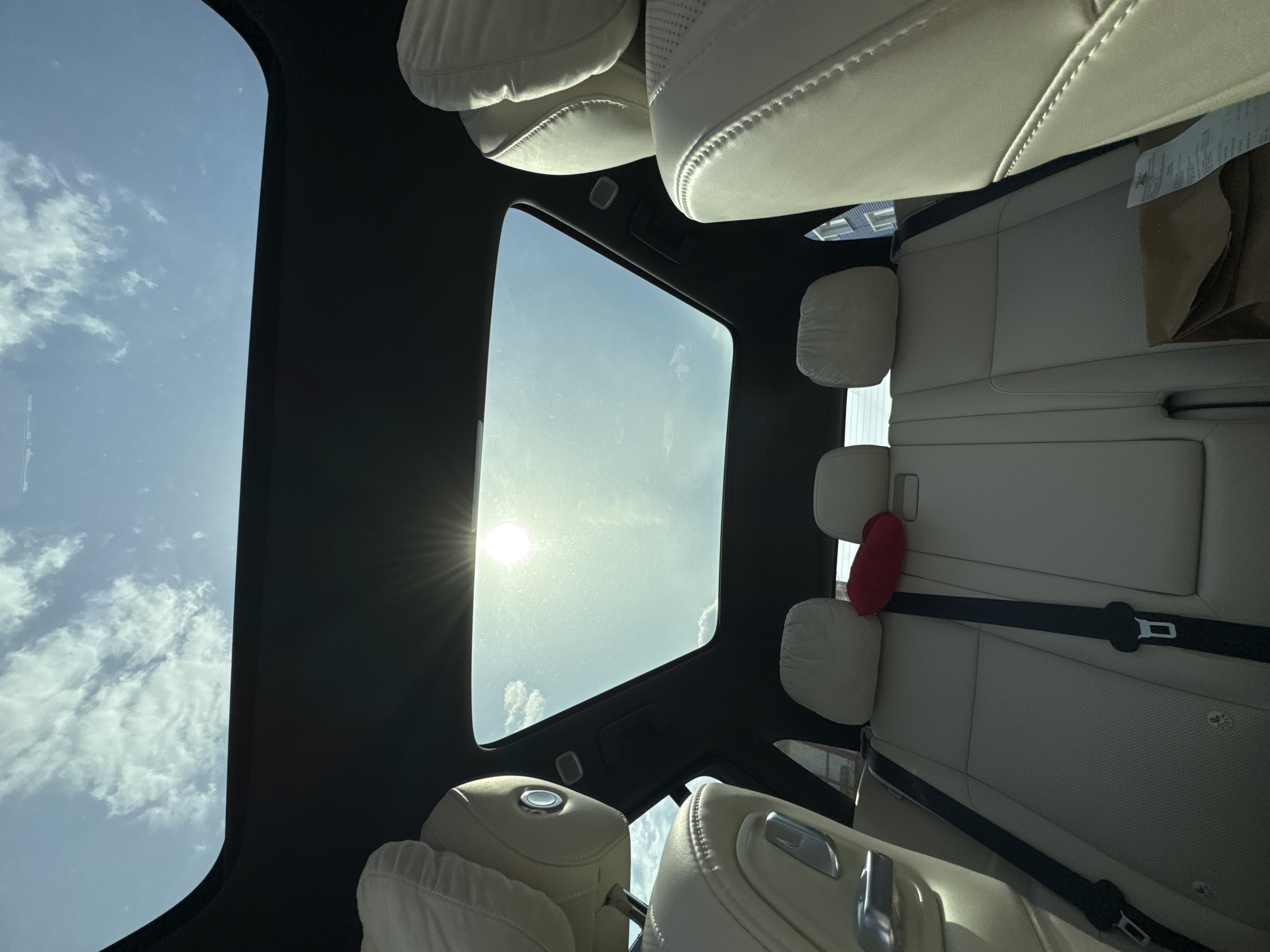 A view looking up through a car sunroof, with the sun shining through the glass. The interior of the car is visible, including the white leather seats and headrests. There is a red object on the back seat. The sky is blue with some clouds visible through the sunroof. Location: Unknown, Unknown.