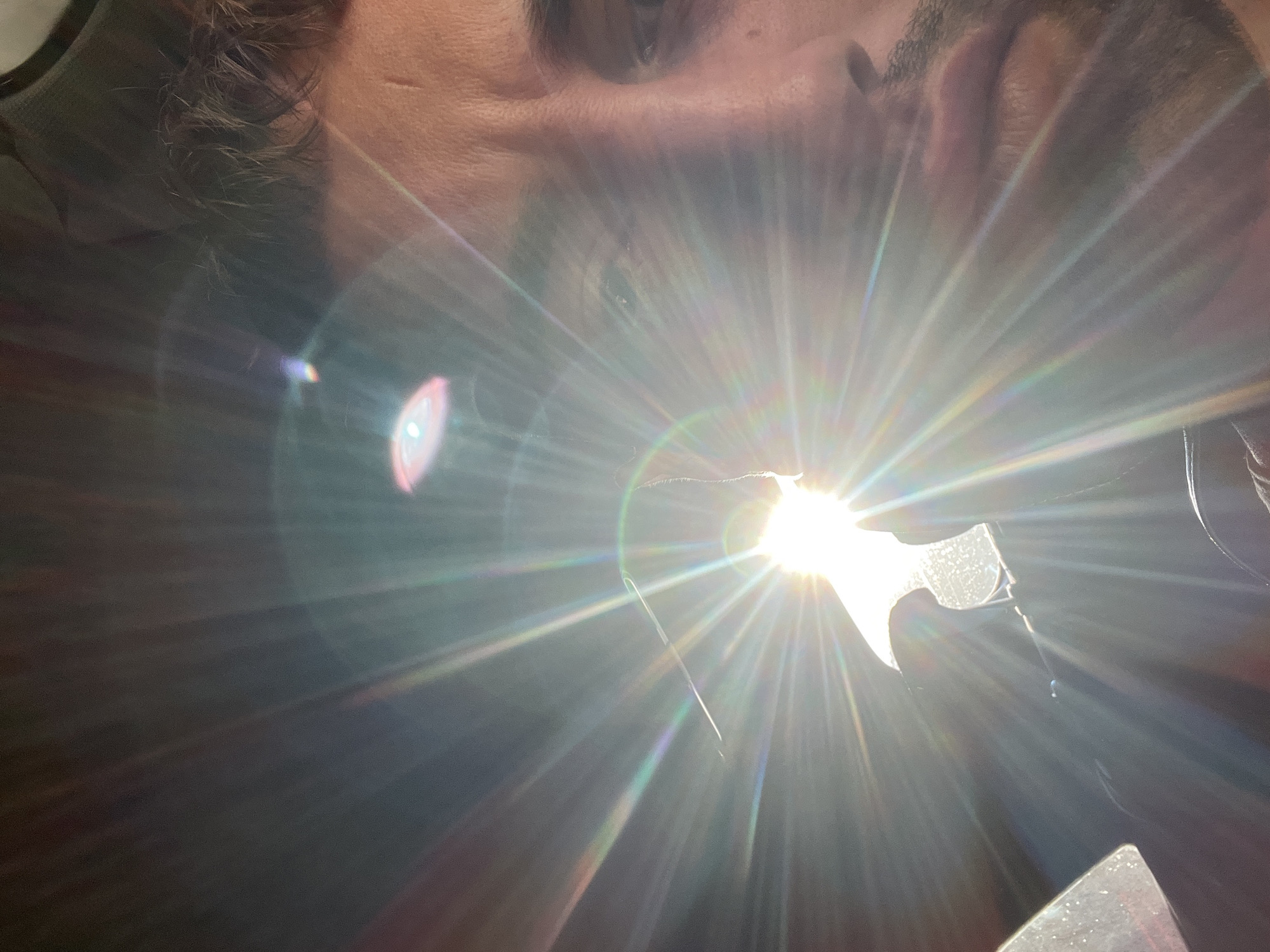 The image is a close-up, overexposed photograph of a man's face, taken from a low angle. The sun is directly in front of the camera, creating a strong lens flare that obscures much of the scene. The man appears to be Caucasian, with short, dark hair and stubble on his face. He is looking towards the camera, and his right eye is visible. The background is dark, with some indistinct shapes that may be part of a car interior. The image does not contain any information about the city of Rogosh, Bulgaria.
