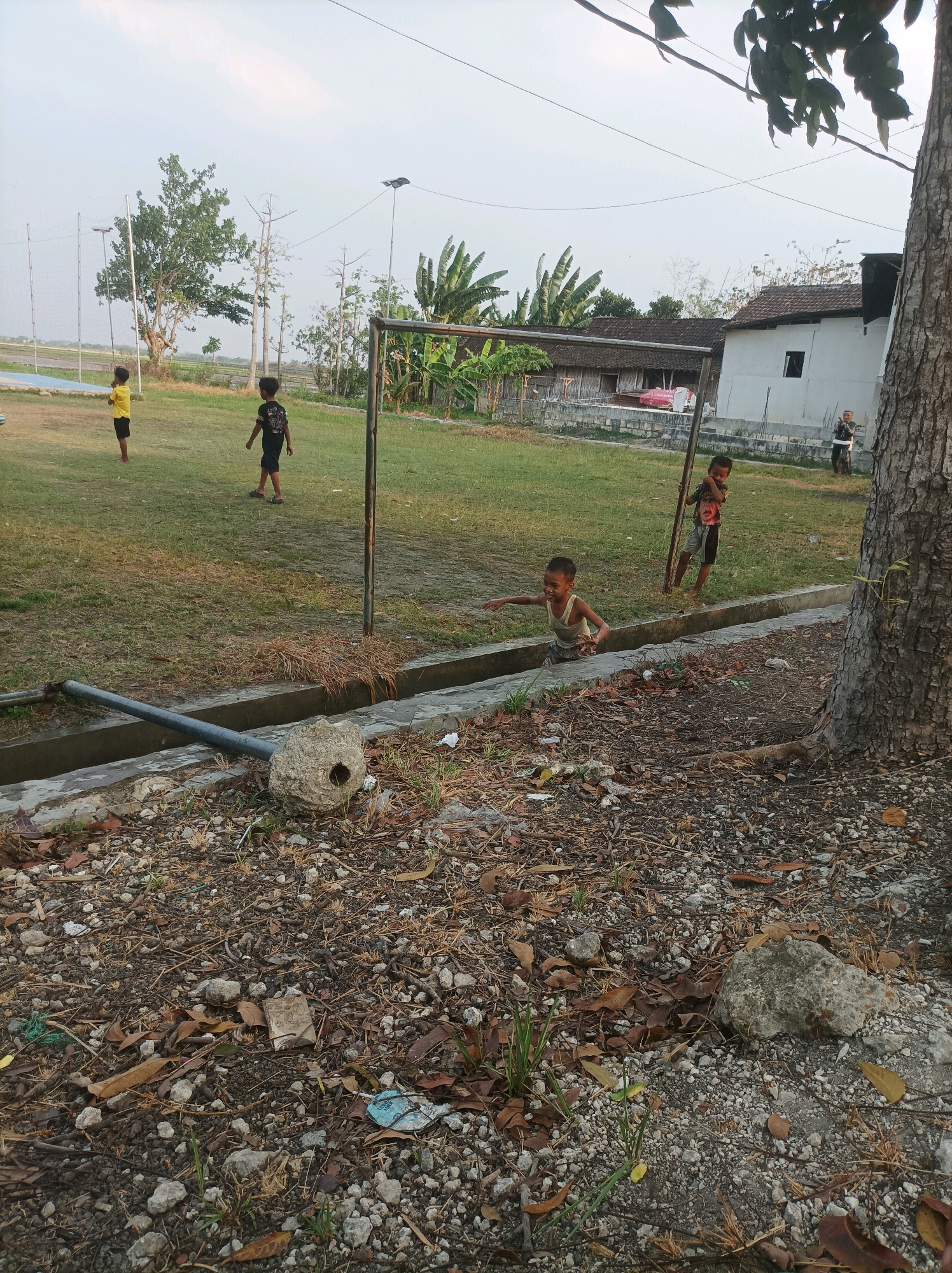 A group of four young boys are playing soccer on a grassy field near a cluster of buildings in Sarangan, Indonesia. The field has a goalpost visible in the center of the frame. The boys are dressed in casual clothing, with two of them wearing t-shirts and shorts. The environment appears to be a rural or semi-rural area, with banana trees and other vegetation visible in the background. The sky is overcast. The ground in the foreground is covered with debris, including leaves, rocks, and some litter. There is a concrete drainage ditch running through the lower part of the image. One boy is in motion, appearing to be kicking or receiving a ball. Another boy stands by the goalpost, while the other two are further away on the field. A large tree trunk is on the right side of the image.