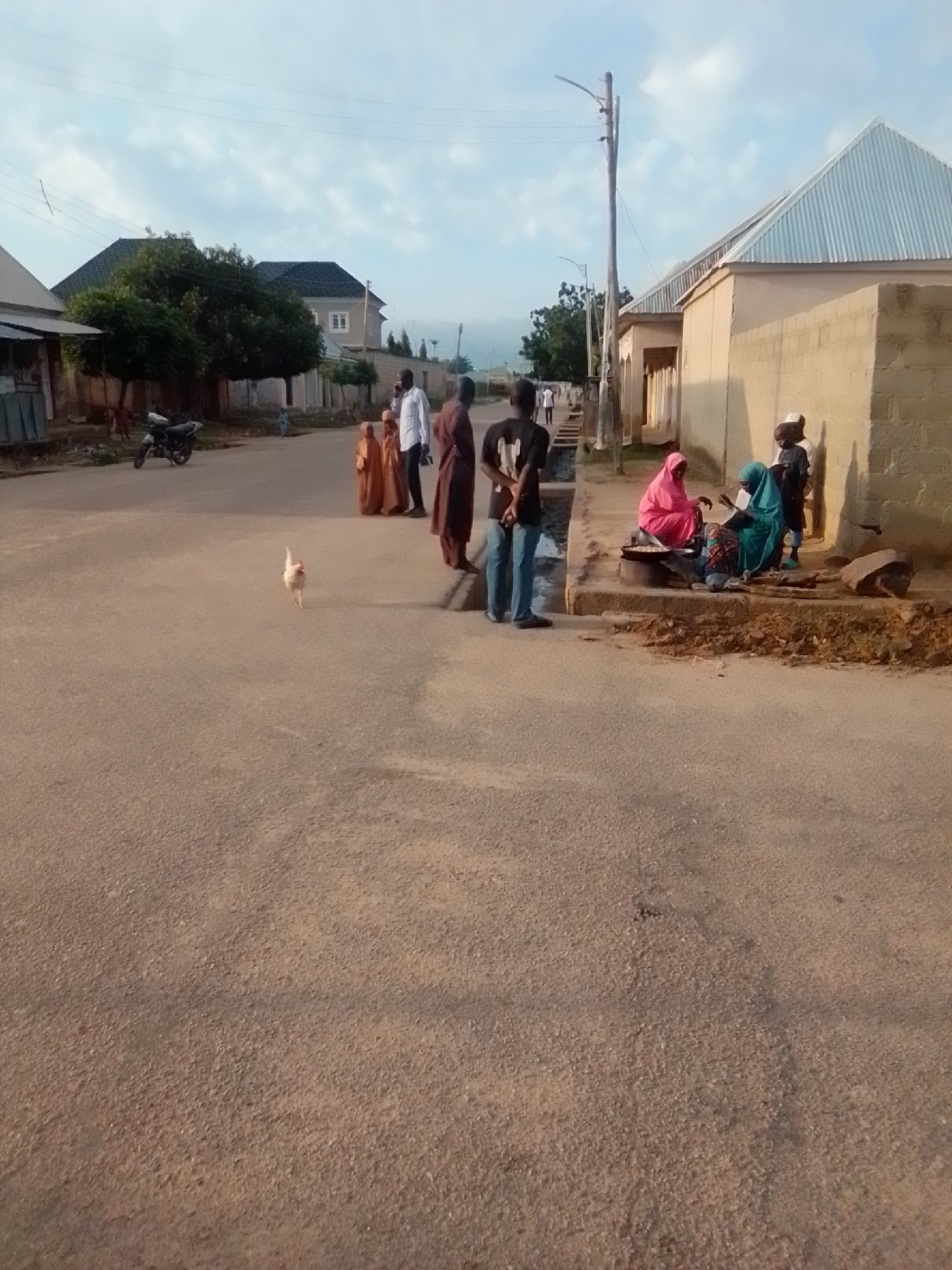 A medium shot shows a paved street in the city of Liji Hill, Nigeria. On the right side of the street, a tan brick building with a silver corrugated metal roof stands tall. A group of individuals are gathered near the building, some sitting on a raised concrete curb. One person is wearing a pink headscarf and appears to be cooking on a portable stove. Another individual wears a teal headscarf and is seated nearby. Other people stand, including a man in a white shirt and a woman in a brown hijab. To the left of the street, a motorcycle is parked in front of a low building with a slanted metal roof. Several trees and bushes are visible behind the buildings, and houses can be seen in the background. The sky is partly cloudy. A chicken walks across the street in the foreground.
