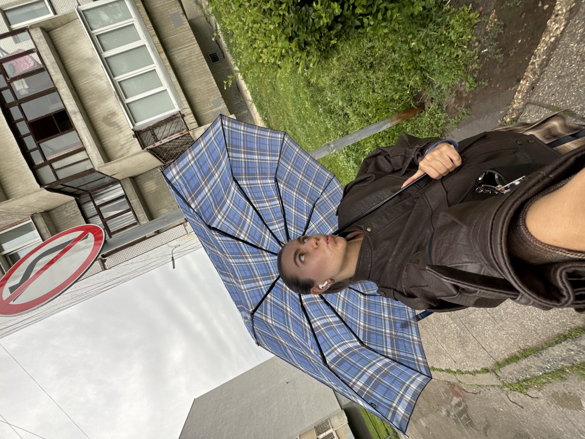 In an urban environment, a young woman stands holding an open, blue and white plaid umbrella, obscuring most of her body. She wears a dark brown leather jacket and what appears to be a plaid skirt underneath. The woman is looking upwards and to her right, with her lips pursed, suggesting she might be taking a selfie. She has white wireless earbuds in her ears. The background shows a multi-story building with balconies and windows, a street sign indicating a "no left turn," and a patch of green grass and bushes. The ground is wet, indicating recent rain. The overall atmosphere is overcast and cool.