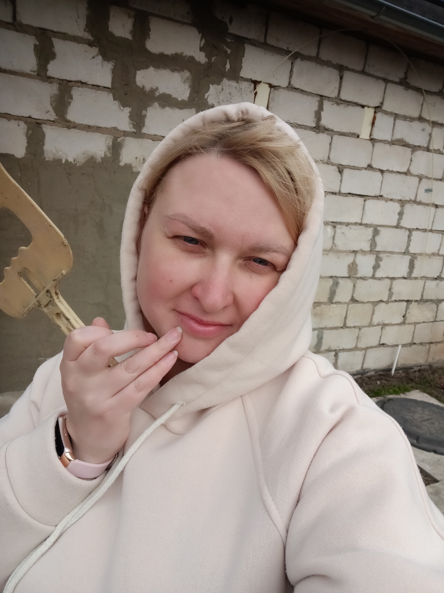 A blonde woman, likely in her mid-20s to 30s, is featured in a close-up outdoor selfie, looking directly at the camera with a subtle, soft smile. She is wearing a light beige or pale pink hooded sweatshirt, with the hood pulled up to cover her head. Her left hand is gently touching her chin and lower lip, revealing light pink fingernails and a pink smartwatch with a rose gold casing on her wrist. On the left side of the frame, partially visible, she holds a cream-colored, somewhat worn plastic object that appears to be a toy or a handle of a tool.
The background consists of a rough, light-colored brick wall with prominent mortar joints and patches of grey concrete or plaster, suggesting an unfinished or weathered building. Below the wall, glimpses of sparse ground cover and a dark manhole cover are visible, indicating an outdoor setting, potentially a backyard, construction site, or less-developed area in Chaikovsky, Russia. The lighting is soft and diffuse, suggesting an overcast day. No discernible text is visible on any subject or in the background.
