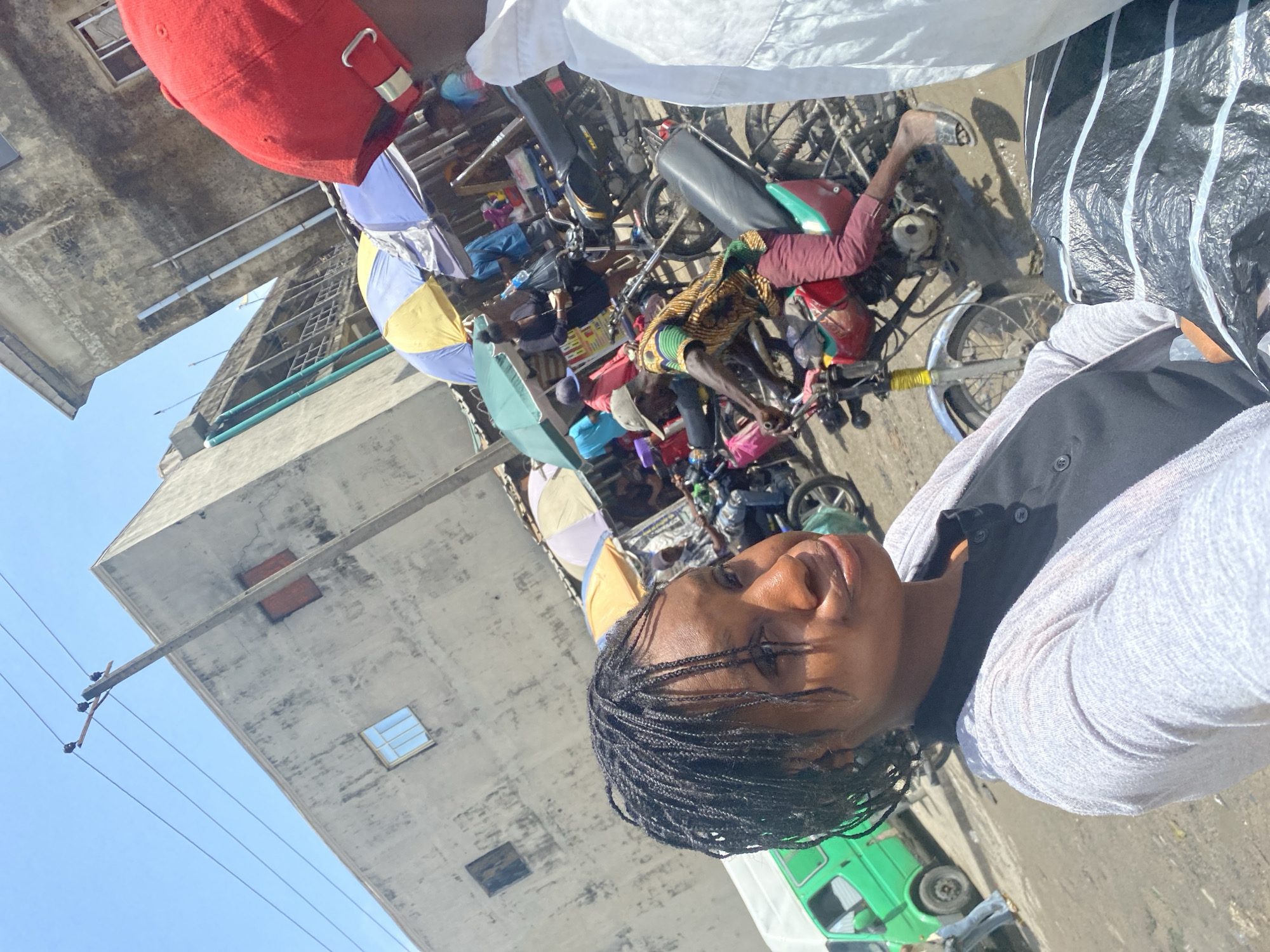 The image captures a vibrant and bustling street scene, likely a market or commercial area, under bright daylight. In the immediate foreground, a person with dark braided hair and dark skin is taking a selfie, looking directly at the camera with a neutral expression. They are wearing a light-colored, possibly white or grey, long-sleeved top over a dark vest. Partially visible above them is another individual wearing a red baseball cap.
The background is a dynamic tableau of daily life. Numerous motorcycles, suggesting a reliance on "okada" (motorcycle taxi) transport, are scattered throughout the scene, some parked and others with people interacting with them. Several market stalls are identifiable by colorful umbrellas—one yellow and blue, another light blue—providing shade for vendors or customers. People are seen milling about, with one individual prominently wearing a yellow, green, and black patterned shirt near a motorcycle. The ground appears uneven, a mix of dirt and possibly some pavement, with patches that might be muddy or dusty.
Multi-story concrete buildings, showing signs of wear and age with visible windows and utility fixtures, line the street under a clear, bright blue sky. Utility poles with wires are also visible, running across the upper part of the image. A green vehicle, possibly a minivan or small bus, is partially visible in the lower right background. The strong sunlight casts clear shadows, indicating it is daytime. No discernible text is clearly visible on any signs or objects. The overall scene suggests a busy urban environment in Satellite Town, Nigeria.