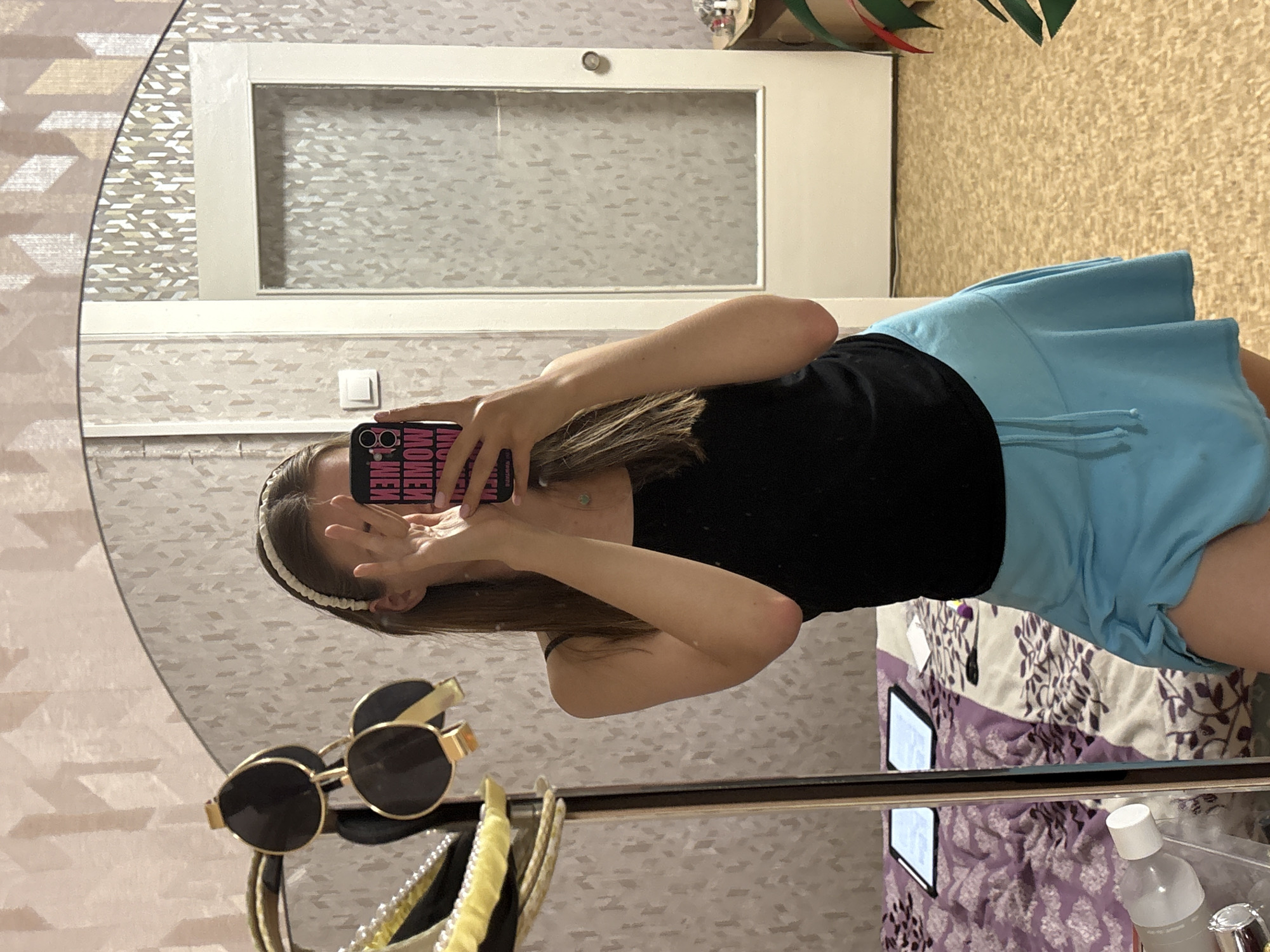 The image shows a woman taking a selfie in a mirror. She is wearing a black top and light blue shorts. The mirror is surrounded by a patterned wall. In the foreground, there are sunglasses and other objects on a shelf or surface. The reflection in the mirror suggests the scene is indoors. Based on visual evidence, I can't verify the location in the city of Perm, Russia.