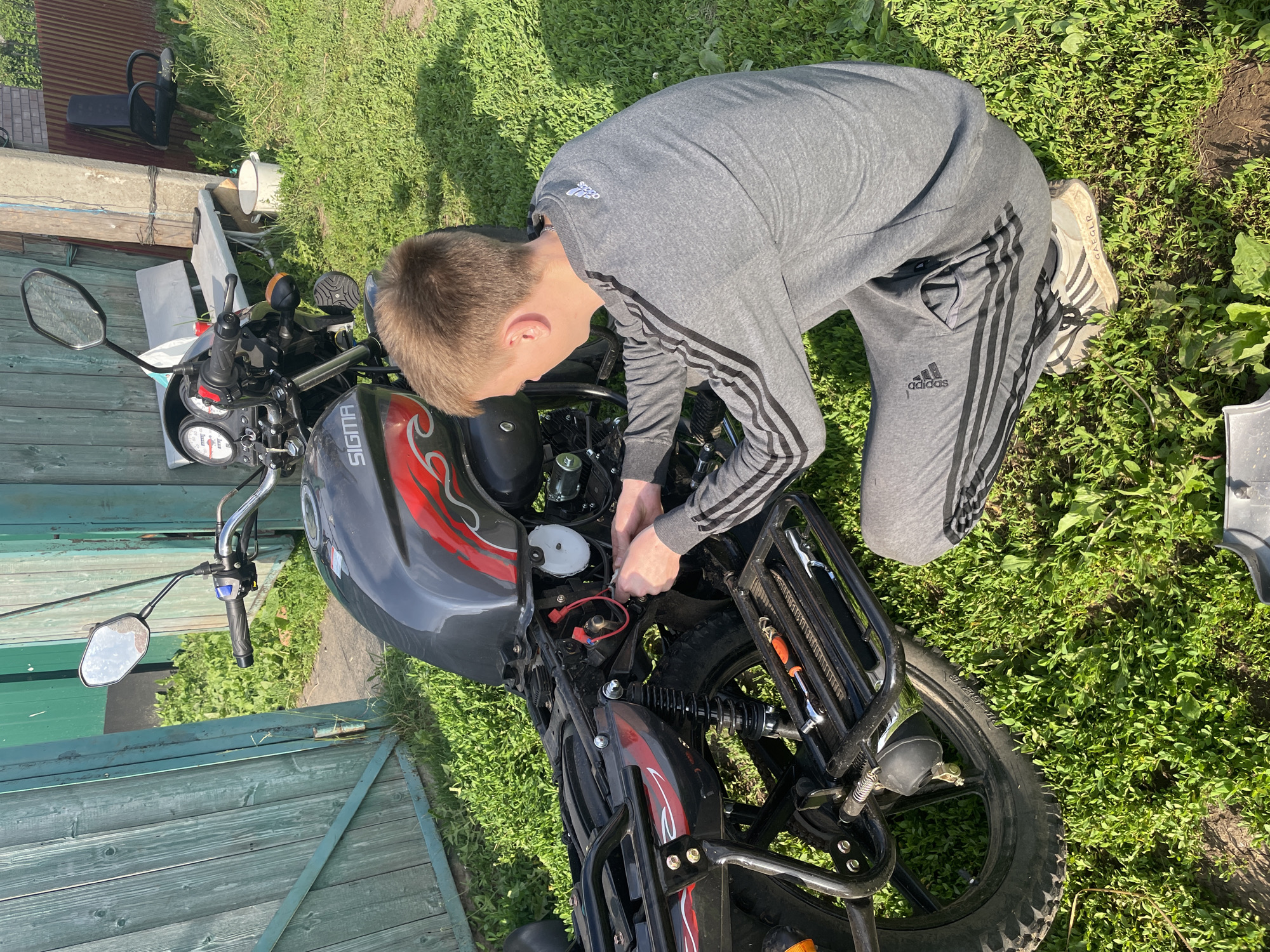 In Nizhnii Bugrysh, Russia, a young male, dressed in a grey Adidas tracksuit and athletic shoes, kneels on a grassy area in front of a motorcycle. He is actively working on the motorcycle's electrical components, his hands engaged with wiring. A toolkit containing an orange-handled screwdriver and a wrench is placed on the motorcycle's rear rack. The background features a weathered green wooden fence and a glimpse of a rural setting with overgrown grass, a bench, a chair, and a white bucket. The scene is illuminated by bright sunlight, casting distinct shadows.