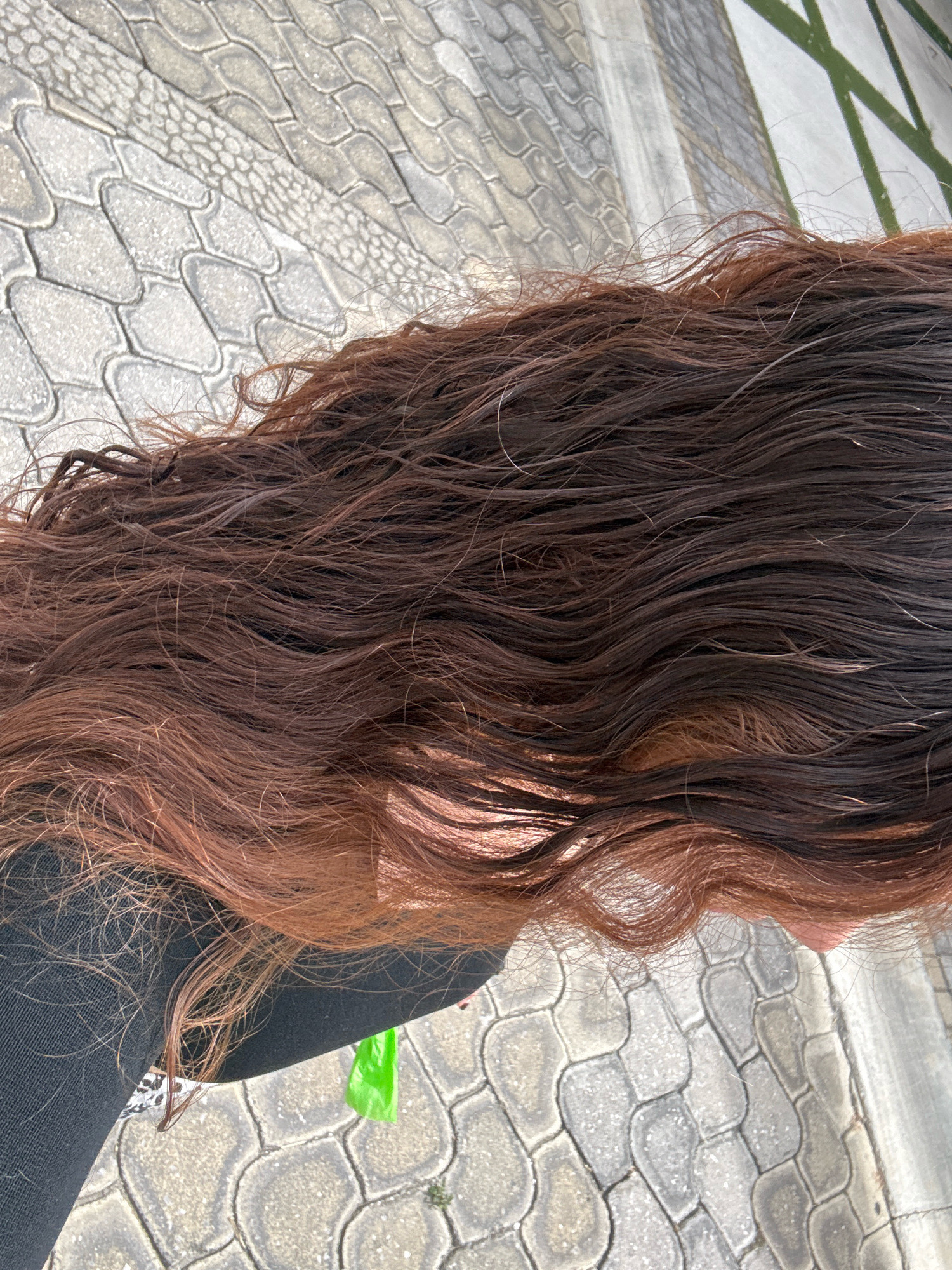 The image shows a close-up of long, wavy, brown hair against a textured gray paver sidewalk. The hair appears to be damp and has varying shades of brown, from lighter reddish-brown to dark brown. A portion of black fabric, likely clothing, is visible on the left side of the image, as well as a bright green plastic bag. The background features a patterned paver ground, typical of outdoor public spaces, and some indistinct green elements that could be foliage or part of a structure. The lighting suggests it is daytime. The location is identified as Santa Clara Ocoyucan, Mexico.