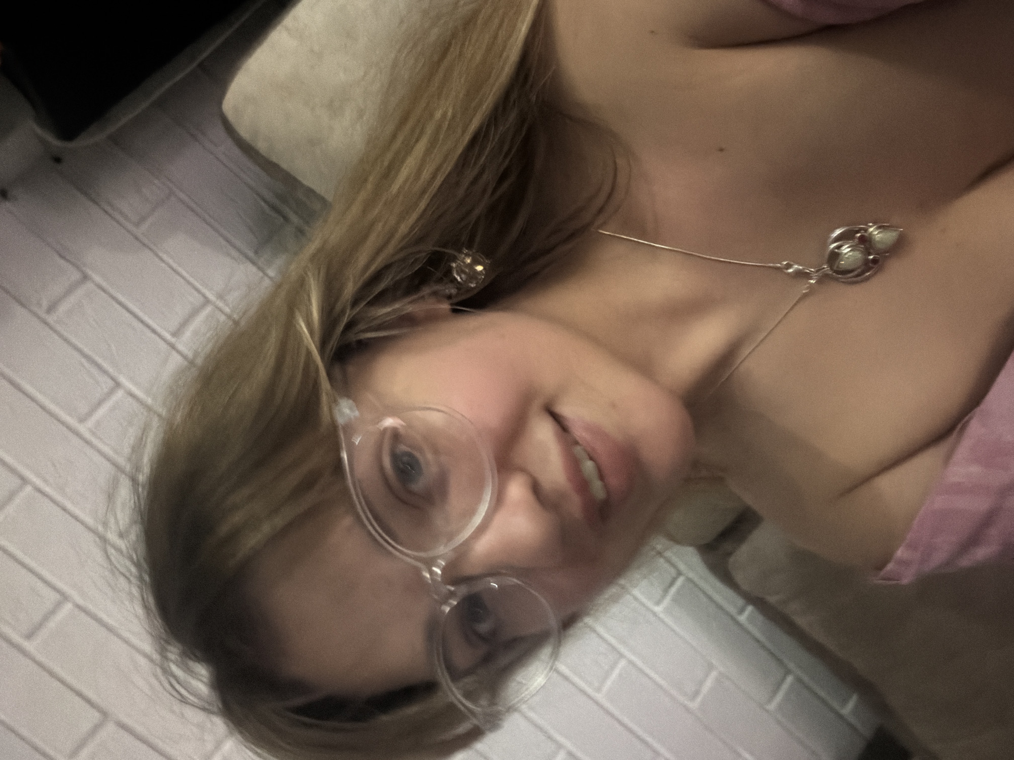 A woman with blonde hair and glasses is photographed indoors. She is wearing a necklace and a pink top. The background includes a white brick wall, a sofa, and what appears to be a black object. The location is Большая Шильна, Russia.