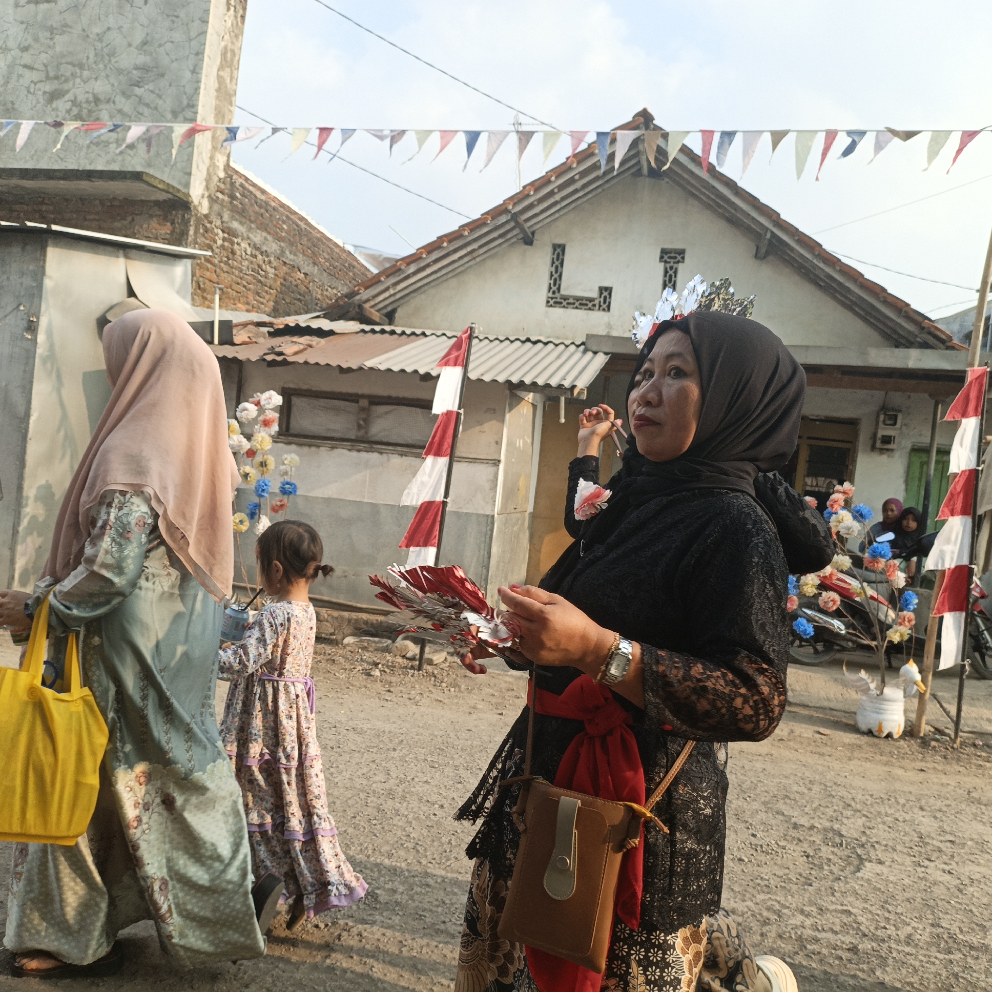 A group of people are gathered on a dusty street in Tanahbaya, Indonesia, under a bright, clear sky. A woman in a black hijab and black lace dress is holding a small, red and white striped sailboat-shaped ornament and a fan-like decoration. She wears a silver crown on her head and a brown crossbody bag hangs from her hip, with a red sash tied around her waist. To her left, a woman in a light pink hijab and a patterned teal and floral dress walks away from the camera, carrying a bright yellow tote bag. Between the two women, a young child in a floral dress with a purple sash stands holding a drink. In the background, a white house with decorative window openings and a red-tiled roof is visible. Red and white triangular flags are strung across the scene, decorating the street. Several motorcycles and a decorative potted plant shaped like a duck are also present in the background. The overall scene appears to be a local celebration or event.