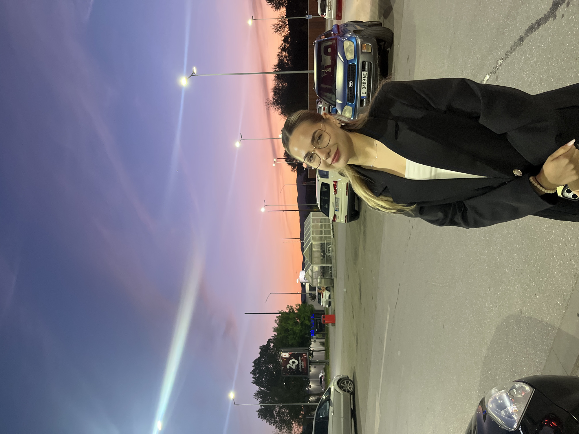 The image depicts a young woman posing in an outdoor parking lot during dusk in Blagoevgrad, Bulgaria. The sky is a prominent feature, showcasing a beautiful gradient from deep purple at the top to soft pink and orange closer to the horizon, indicative of twilight or sunset.
The main subject is a light-skinned woman, likely with light brown or blonde hair, wearing glasses with clear frames. She is dressed in a black blazer over a white top, accessorized with a delicate necklace and a beaded bracelet on her right wrist. Her nails appear to be light-colored. She looks towards the camera with a slight, pleasant smile.
The background reveals a paved parking area with several parked cars. A blue SUV with the Bulgarian license plate "E 0489 MC" is clearly visible behind the woman. Another white car in the mid-ground displays the license plate "CA 8380 AB". Various streetlights are illuminated throughout the scene, casting beams of light, particularly from the upper left. Trees line the background, and a structure resembling a car wash or commercial canopy is visible further back. A dark sign showing "20" is partially obscured by trees. The overall setting suggests a commercial complex or a large retail area.