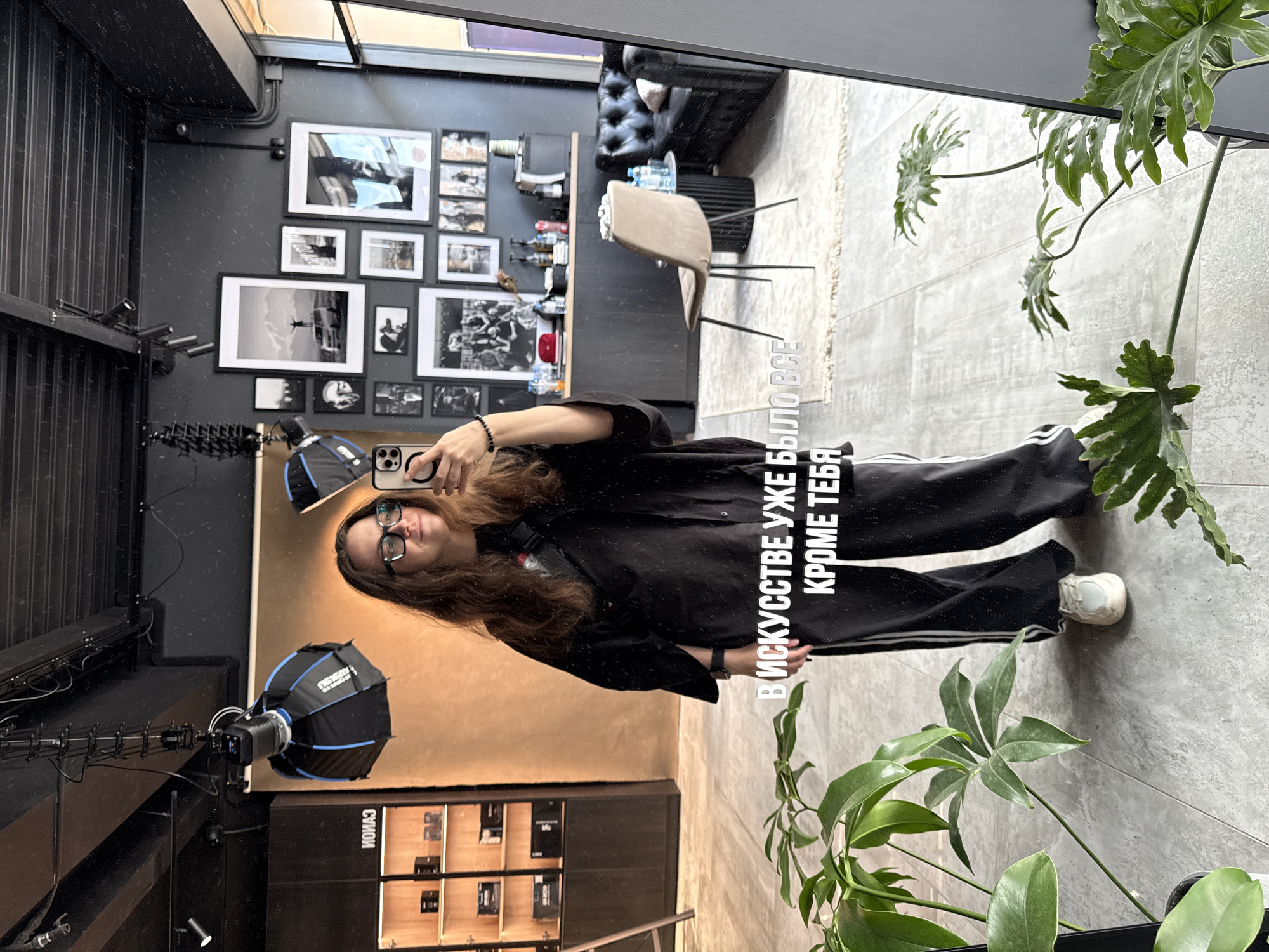 A mirror selfie shows a woman in a black tracksuit, wearing glasses, holding an iPhone. The reflection reveals a room with a black and tan color scheme. On the black wall are framed black and white photographs. Also reflected are a black leather sofa, a tan-colored chair, and various items on shelves. Photography equipment, including a strobe with a softbox, is visible. A potted plant sits near the mirror's edge. Text in Cyrillic is superimposed on the mirror, partially obscuring the image. The location is identified as Nizhny Novgorod, Russia, based on a previous instruction.