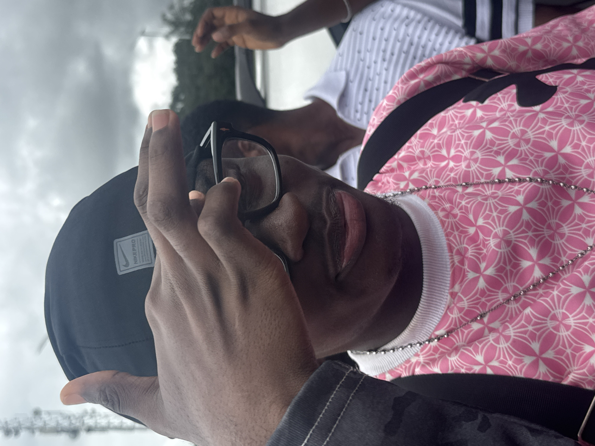 A close-up, medium shot captures a dark-skinned individual from the chest up, wearing a black Nike Pro cap and glasses. Their right hand is raised, obscuring their forehead and left eye. They are wearing a pink t-shirt with a white geometric pattern and a silver chain. A person in a white polo shirt is visible in the background. The scene appears to be outdoors, with a cloudy sky and trees. The image shows a communication tower in the distance, which is a prominent landmark in Ihiagwa, Nigeria, suggesting the photo was taken in that location.