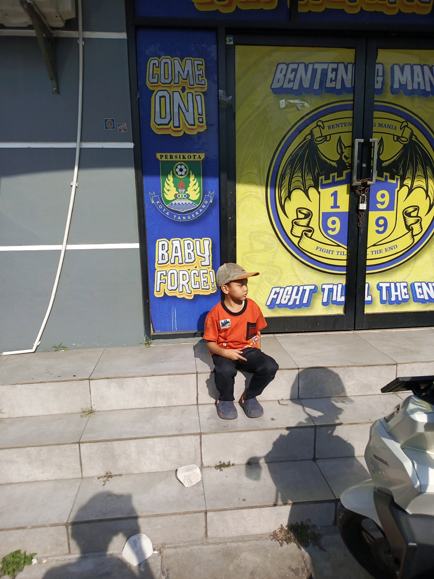 A young boy wearing an orange t-shirt, black pants, a baseball cap, and grey crocs sits on concrete steps in front of a building with blue and yellow signage. The signage on the left reads "COME ON!" and features a crest for "PERSIPOTTA KOTA TANGERANG". The signage on the right has a yellow background with a crest featuring a bat and the text "BENTENG MANIA 1999". Below this crest, it reads "FIGHT TIL THE END". The boy is looking to his left. A motorcycle is partially visible in the lower right corner of the image. The scene appears to be outdoors in daylight.