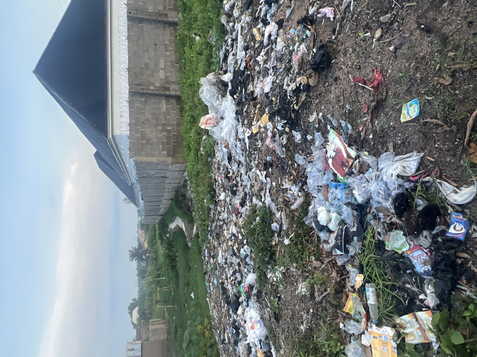 The image depicts a large pile of garbage in an outdoor setting. The garbage consists of various items, including plastic bags, bottles, wrappers, and discarded food packaging. Some of the items are colored, while others are transparent. The ground is covered with dirt and debris, with some patches of green grass growing through the trash. In the background, there is a wall constructed of concrete blocks, topped with barbed wire. Beyond the wall, a building with a dark, pitched roof is visible, along with trees and a cloudy sky. The scene appears to be located in a somewhat developed area, possibly on the outskirts of a city.
