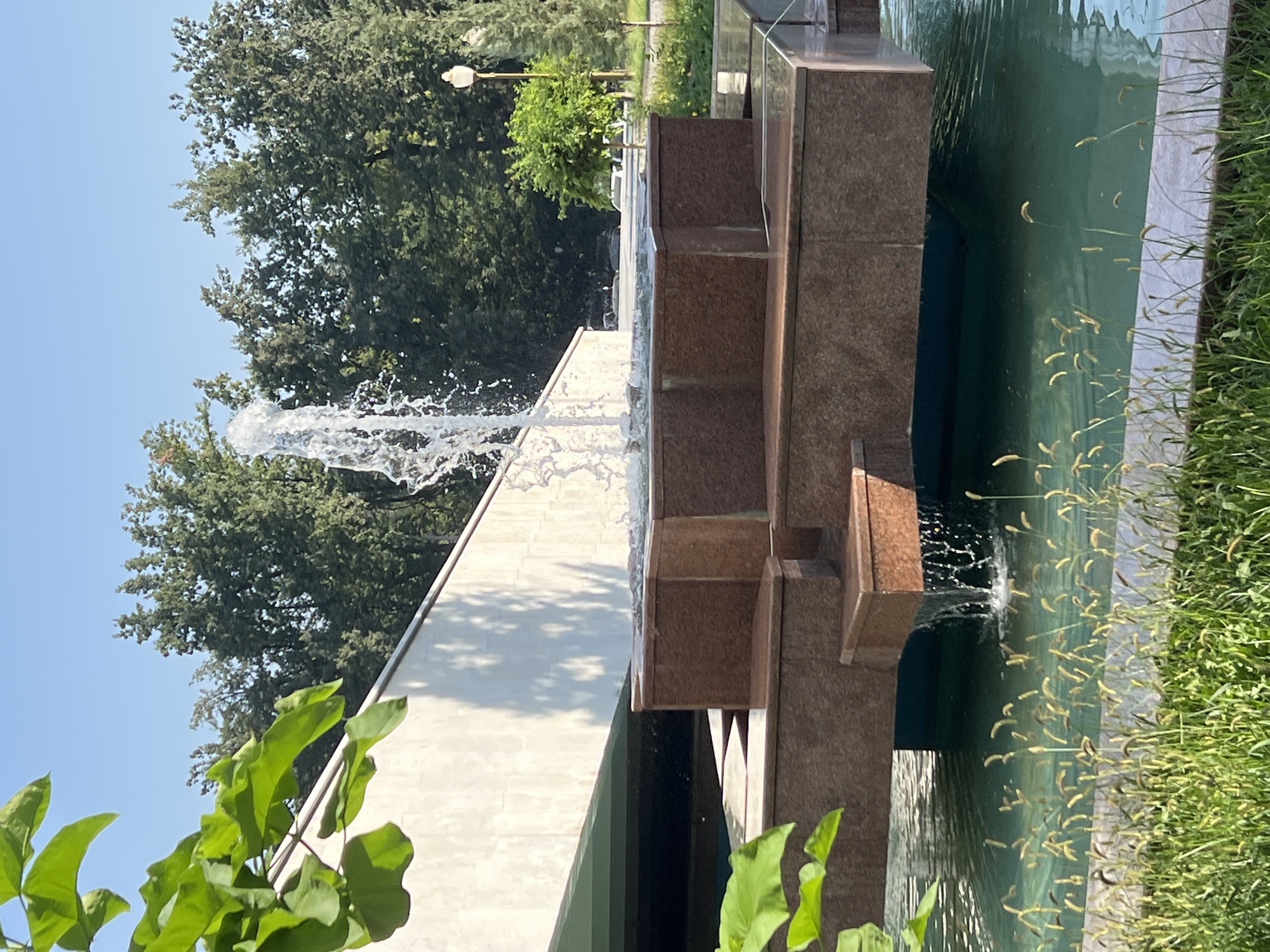 The image depicts a tiered fountain with water cascading down its levels into a pool. The fountain is constructed with brown granite and a teal-colored base. In the background, a white, angled wall with visible shadows is present, leading up to a line of lush green trees under a clear blue sky. To the right, a lamppost and a small green tree are visible, along with more trees and foliage in the distance. The foreground features green grass and tall, thin plants with seed heads growing in front of the fountain's pool. No people are visible in the frame. The scene appears to be an outdoor public space, possibly a park or plaza.