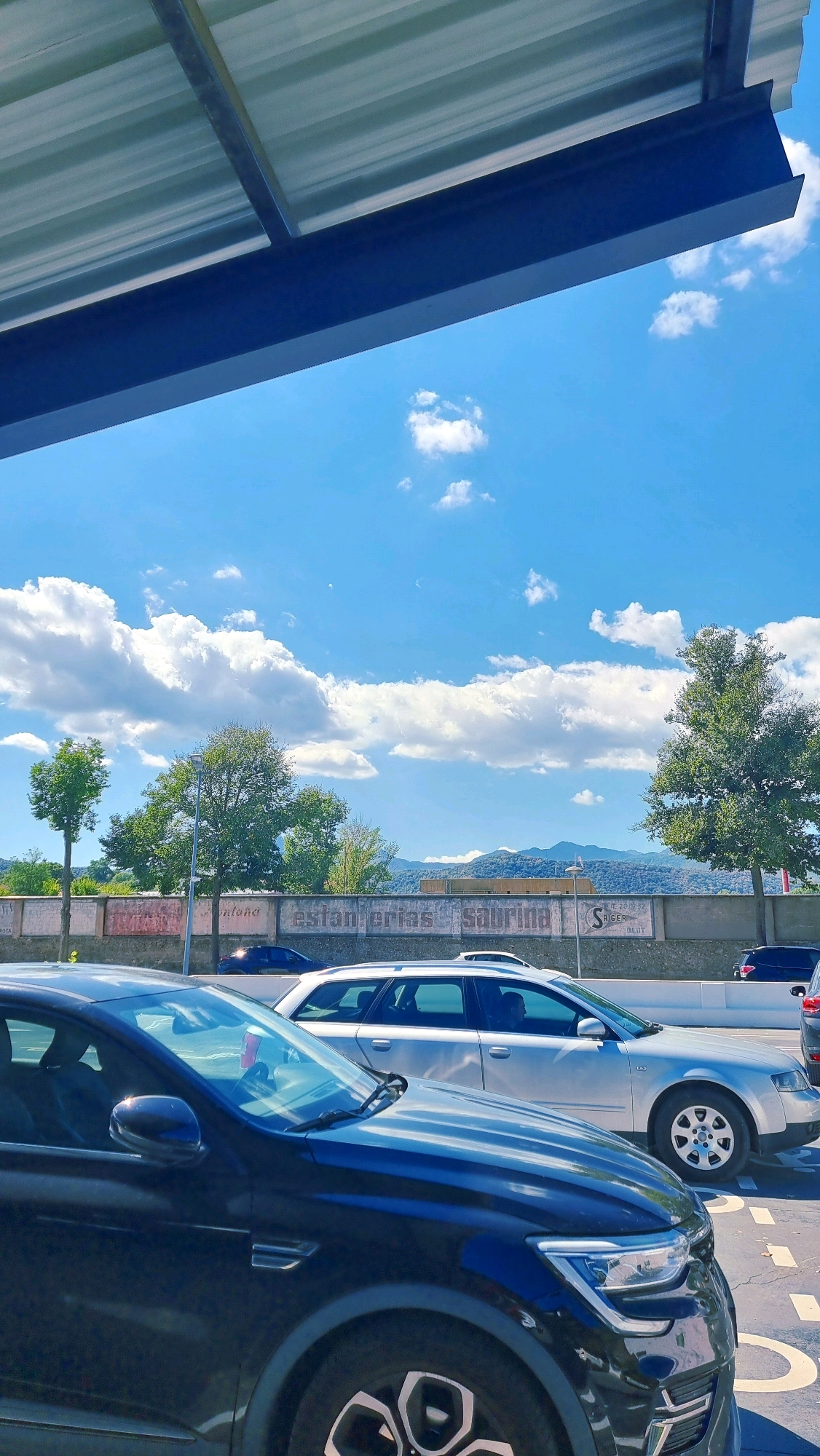 The image shows a street scene in Olot, Spain, with cars and a building in the background. The sky is blue with some white clouds. A dark blue car is in the foreground, and a silver station wagon is behind it. In the background, there is a building with some writing on it that says "ESTANTERIAS SABRINA". There are also some trees and mountains in the distance. The scene appears to be a sunny day.