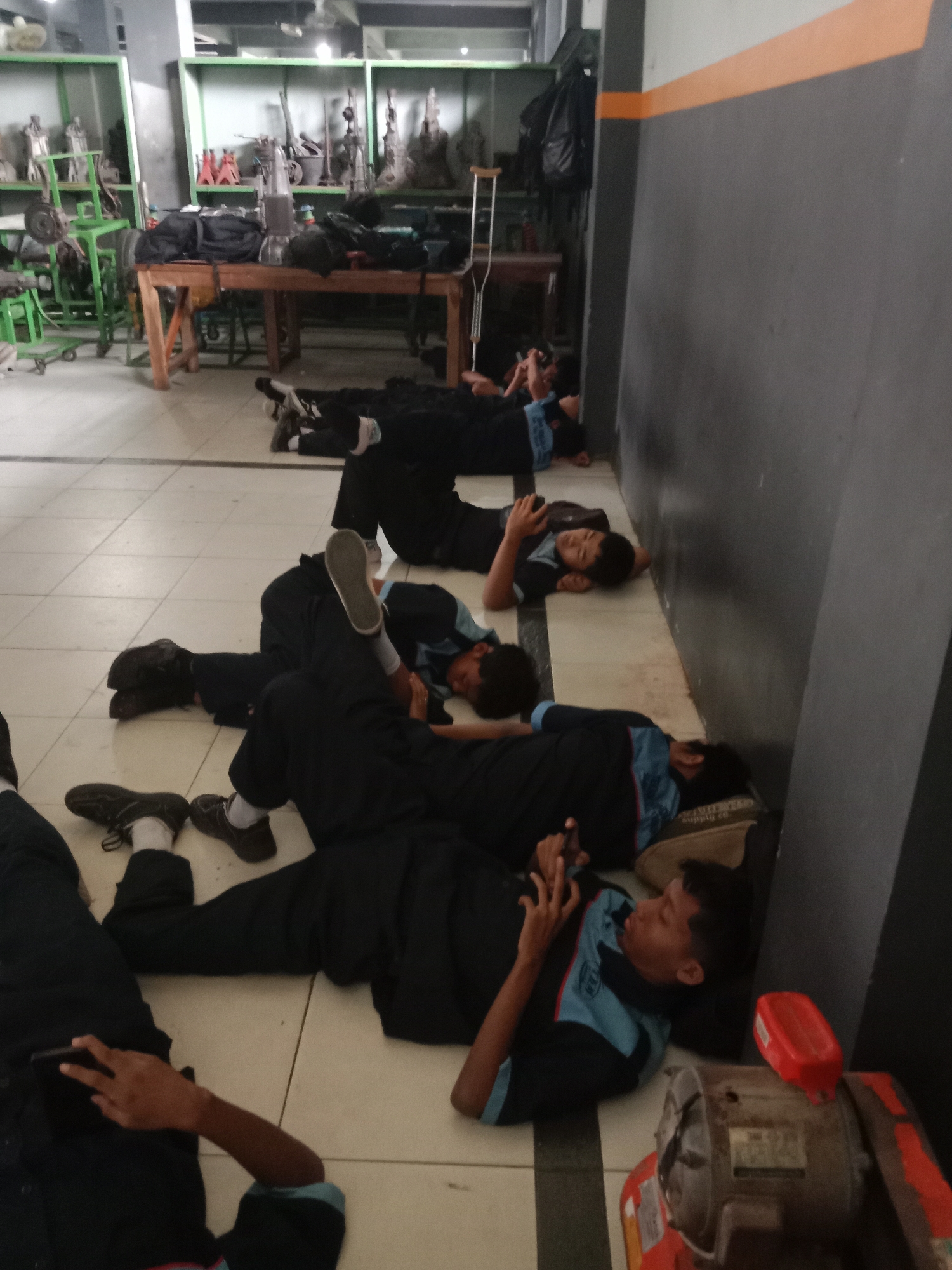 In a workshop environment in Kebumen, Indonesia, multiple young males are observed lying on the floor, appearing to be sleeping or resting. They are dressed in what seem to be uniforms, characterized by dark trousers and light blue shirts with darker blue accents on the collars and sleeves. Many of the individuals are holding or looking at mobile phones, suggesting a moment of leisure or a break. The room contains various objects indicative of a workshop or vocational training space, including tables laden with tools and equipment, metal shelving units displaying more tools and parts, and a large machine with a red component in the foreground. The lighting suggests an indoor setting, possibly under fluorescent lights, with a gray and orange striped wall in the background. The floor is tiled with a checkerboard pattern.