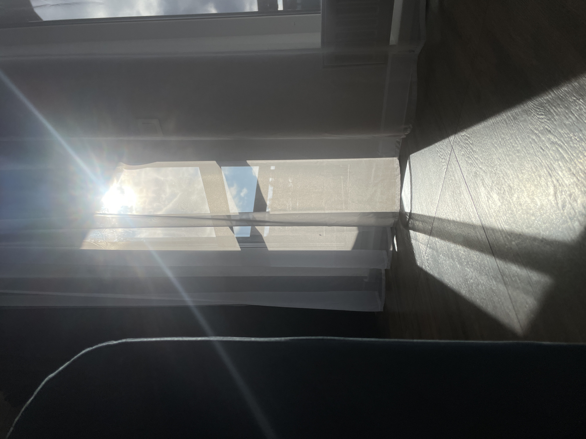 The image captures an indoor scene dominated by bright sunlight streaming through a window. The original image is oriented vertically. On the left side, strong sun rays penetrate the room, creating a prominent lens flare effect near the center-left. A window with sheer white curtains or blinds is visible, allowing significant light to pass through and illuminating the textures of the fabric. Between the curtain panels, a patch of blue sky with white clouds is visible, suggesting a clear day outdoors.
To the right of the window, a wooden or laminate floor is brightly lit by the direct sunlight, showcasing its grain and texture. Long, distinct shadows are cast across the floor, likely from the window frame and interior elements, indicating the low angle of the sun, possibly morning or late afternoon. The foreground, occupying the bottom portion of the image, features a dark, blurry object, likely part of a sofa or bed, suggesting the perspective is from a seated or reclining position.
There are no people or specific activities depicted. The scene is static, focusing on the interplay of light and shadow within the room. No visible text is present. The bright, direct sunlight suggests a sunny day. This snapshot captures a tranquil moment within a room in Podshivalovo, Russia, emphasizing natural light.