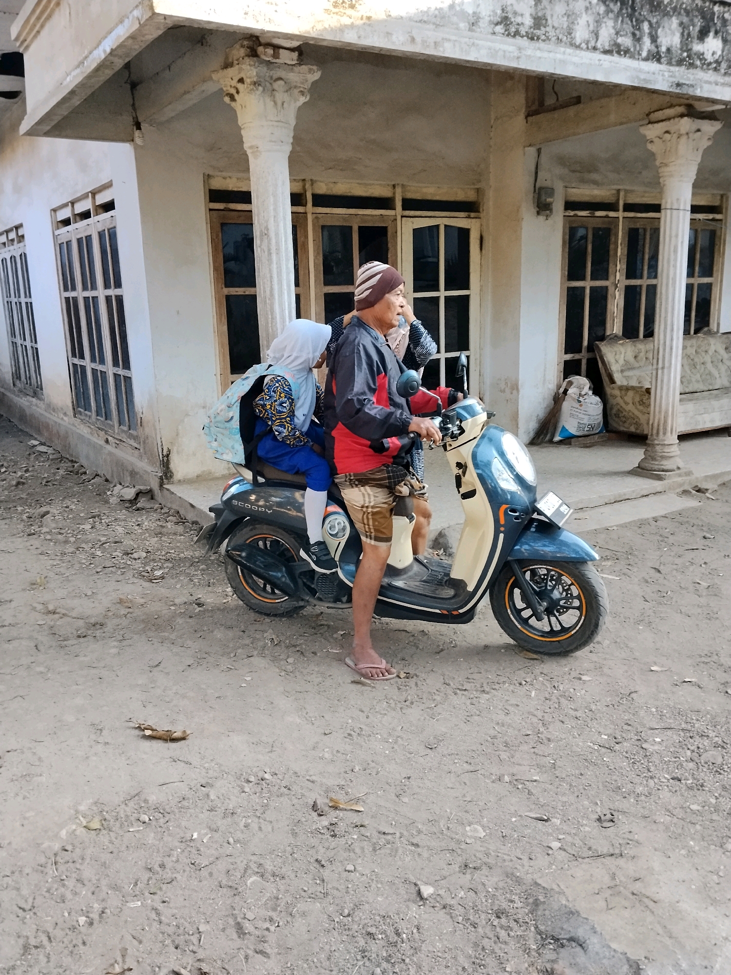 An elderly man wearing a beanie and a patterned jacket is riding a dark blue and cream colored scooter. He is looking to his right and appears to be speaking. A young girl wearing a hijab and blue pants is sitting behind him, facing forward. She has a backpack on and is holding onto the man. The scooter is parked on a dirt and gravel road in front of a weathered, two-story building with white walls and wooden windows. There are white columns supporting an overhang. The building appears to be in a state of disrepair, with cracks in the concrete and peeling paint. The area is generally dusty and unkempt, with scattered debris on the ground. The scene is consistent with a residential street in Tlogosari, Indonesia.
