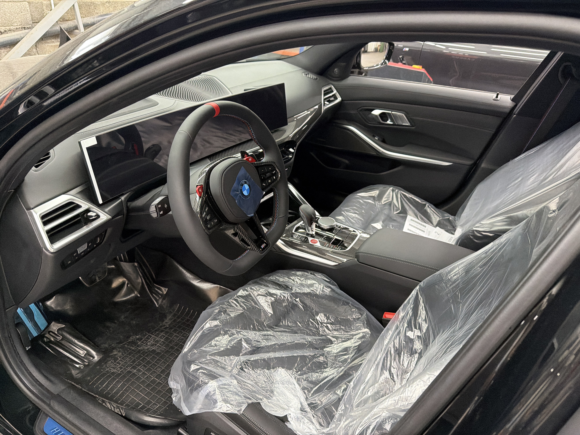 The image shows the interior of a black car, likely a BMW, with the driver's side door open. The seats and steering wheel are covered in protective plastic, indicating the car is new or being prepared for sale. The dashboard features a large screen and carbon fiber accents on the steering wheel. A red and black steering wheel with paddle shifters is visible. The floor has black rubber mats. The car appears to be in a showroom or dealership environment, possibly in Burgas, Bulgaria, suggested by a partial blue and white sign with "Burgas" text visible on the door sill. No people are present in the image.