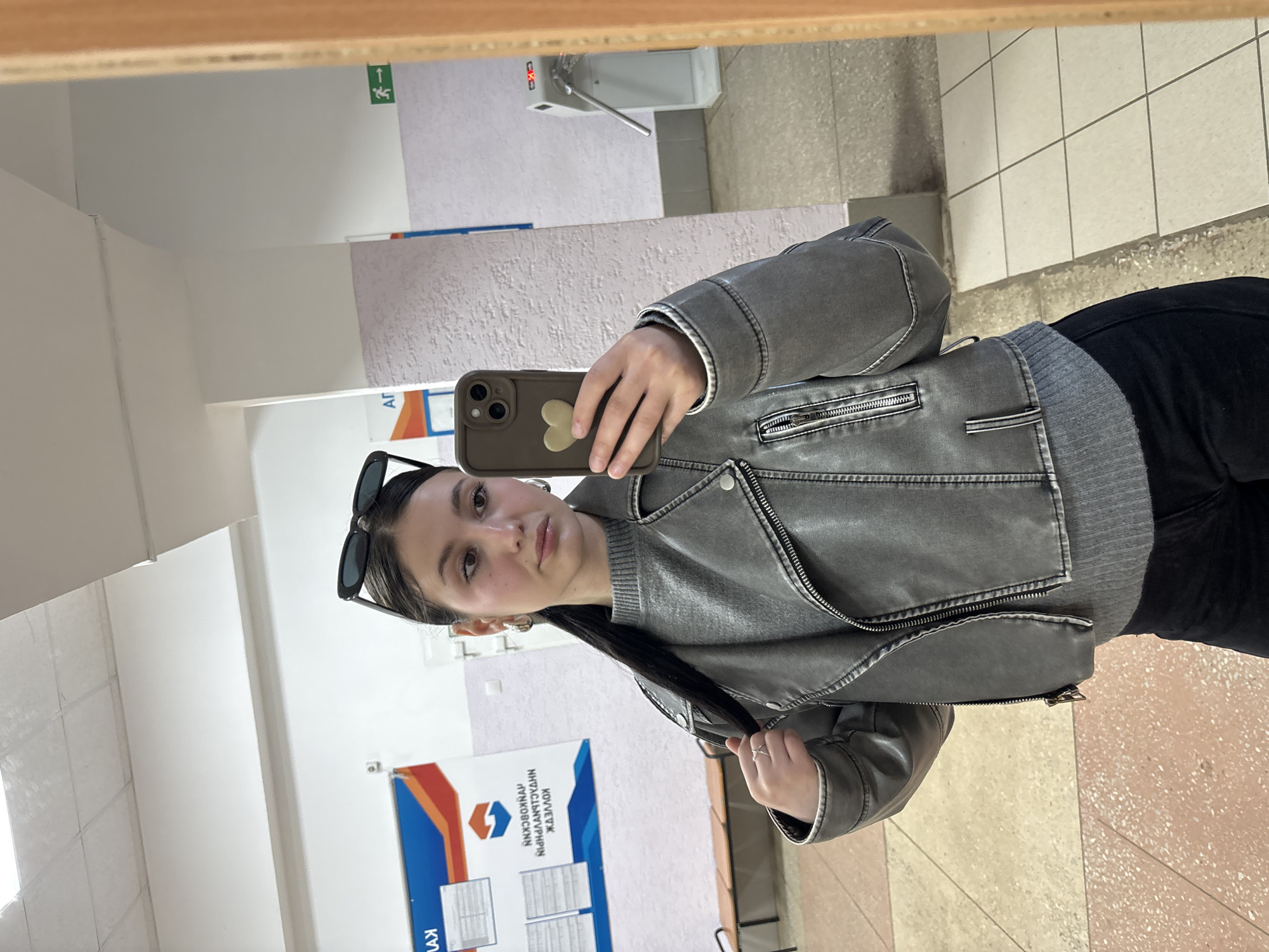 A young woman is captured in a mirror selfie, standing in what appears to be an indoor hallway. She is positioned towards the center, holding a smartphone with a light brown case and a heart-shaped detail in her right hand. Her dark hair is styled back, and black sunglasses are pushed up onto her head. She wears a grey faux leather jacket, detailed with zippers and ribbed knit cuffs and hem, layered over a grey sweater and dark trousers.
The reflected background reveals a bright, institutional-style setting. The walls are light-colored, some with a textured finish, and the floor is tiled with a pattern of light and reddish-brown speckled squares. In the upper left of the reflection, a green "exit" sign with an arrow pointing right is visible. To the upper right, a white utility fixture, possibly a water cooler or sink, is reflected.
A prominent blue and orange banner or sign is displayed on the wall behind her. Visible Cyrillic text on this banner includes "КОЛЛЕДЖ" (College), "КУНГУРСКИЙ" (Kungur), "МУЗЫКАЛЬНЫЙ" (Musical), "ПЕДАГОГИЧЕСКИЙ" (Pedagogical), "ИМЕНИ" (named after), and "ЖДАНОВА" (Zhdanov). This text suggests the location is the "Kungur Musical Pedagogical College named after Zhdanov." Other partial Cyrillic letters like "АГ" and "КУУ" are also discernible on adjacent papers or signs. The indoor lighting suggests artificial illumination, and the woman maintains a neutral expression.