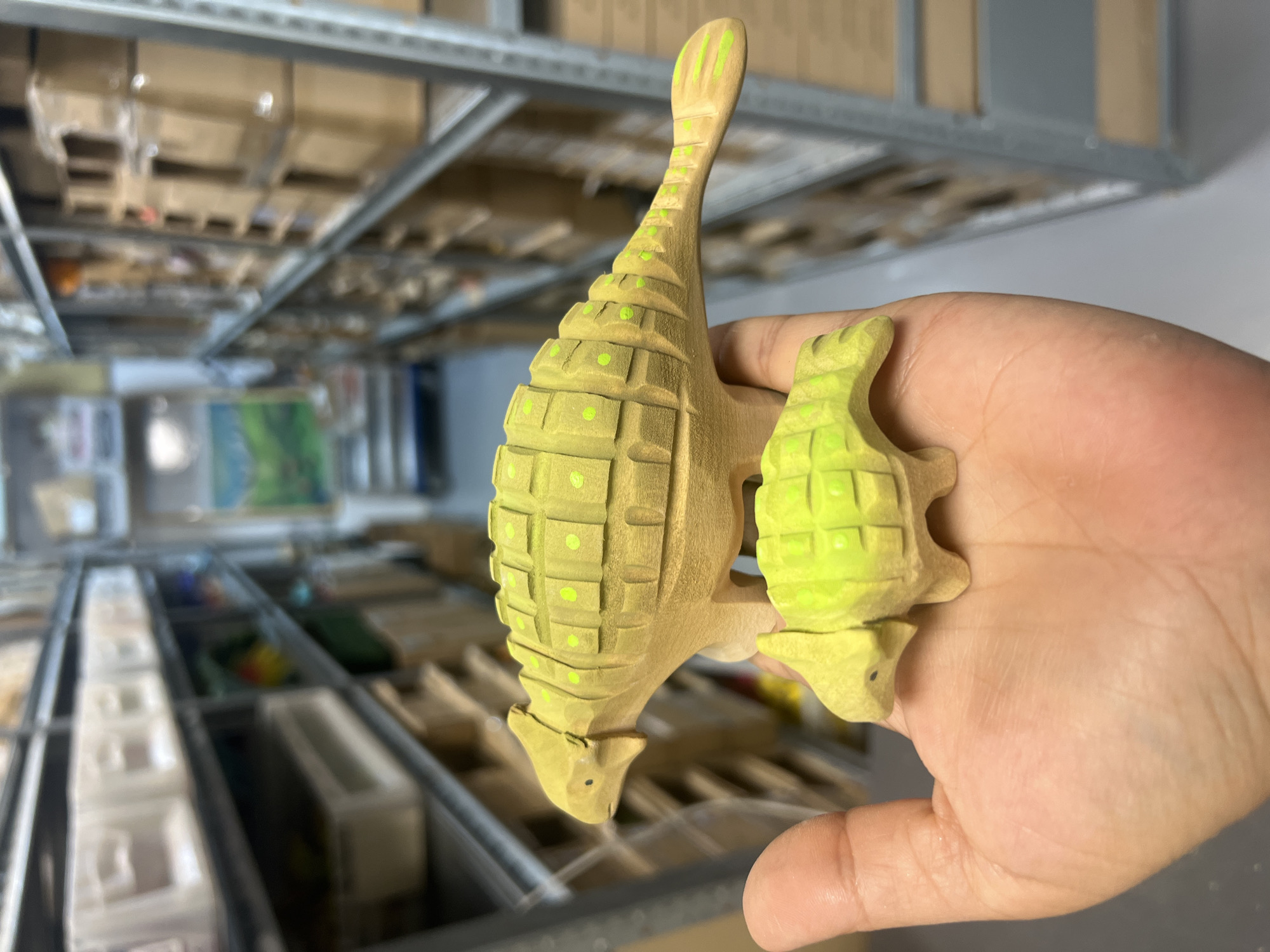 A close-up shot captures a hand holding two wooden dinosaur toys, likely ankylosaurs, in a warehouse setting. The larger dinosaur is positioned on the palm, facing left, its back adorned with carved squares and green dots. A smaller, similarly designed dinosaur rests on top of the larger one, also facing left. The background is blurred but shows shelves filled with boxes and bins, characteristic of a storage facility. The lighting is even, highlighting the texture and detail of the toys. The overall scene suggests a moment of handling or display of these handcrafted items within a commercial or industrial space.