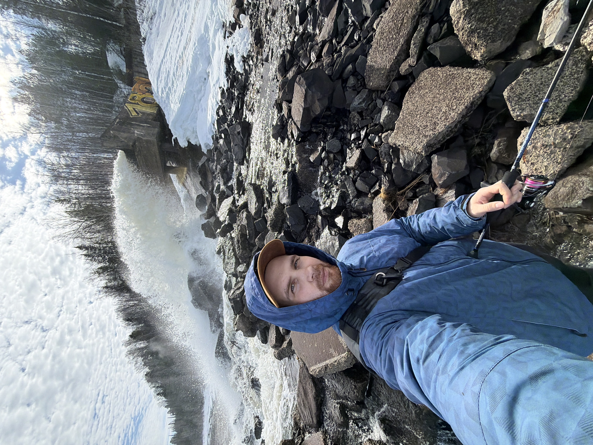 The image depicts an outdoor scene in the city of Unknown, Unknown, at a location characterized by a powerful water discharge and rocky terrain during daylight hours.
A single male individual, Caucasian with a short reddish-brown beard, is centered in the foreground, facing the camera with a neutral expression, appearing to take a selfie. He wears a blue patterned hooded jacket with the hood up, covering a brown baseball cap worn backward. A dark strap crosses his chest. In his right hand, he holds a black and blue fishing rod with a pink and black reel.
Behind the individual, turbulent white water is forcefully discharged from a grey concrete structure into a rocky channel. The concrete structure exhibits yellow graffiti "7850" on its visible side. To the right of the discharge, a large accumulation of snow and ice covers the ground. The immediate foreground and midground consist of dark, uneven rocks and broken concrete slabs, many appearing wet from water spray.
A dense forest of both evergreen and bare deciduous trees lines the horizon. The sky is largely covered by patchy white clouds, with bright sunlight visible in the upper right quadrant.