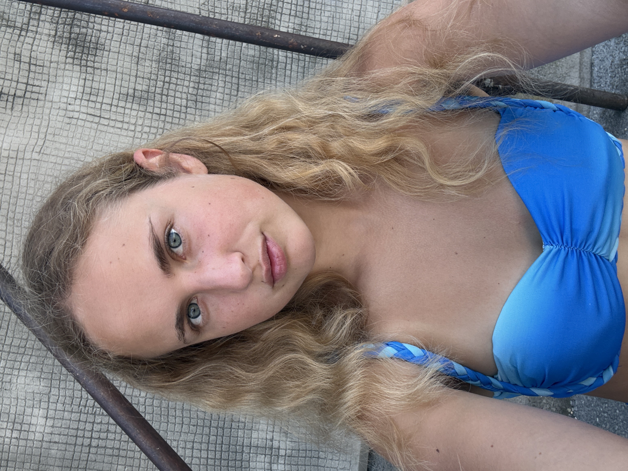 A young woman with blonde, wavy hair and blue eyes is posing for a selfie outdoors. She is wearing a blue bikini with a braided strap. The background appears to be a textured wall and metal bars, possibly part of an outdoor seating area or railing. The lighting suggests it is daytime. The location of Trieste, Italy, is not discernible from the visual information in the image.