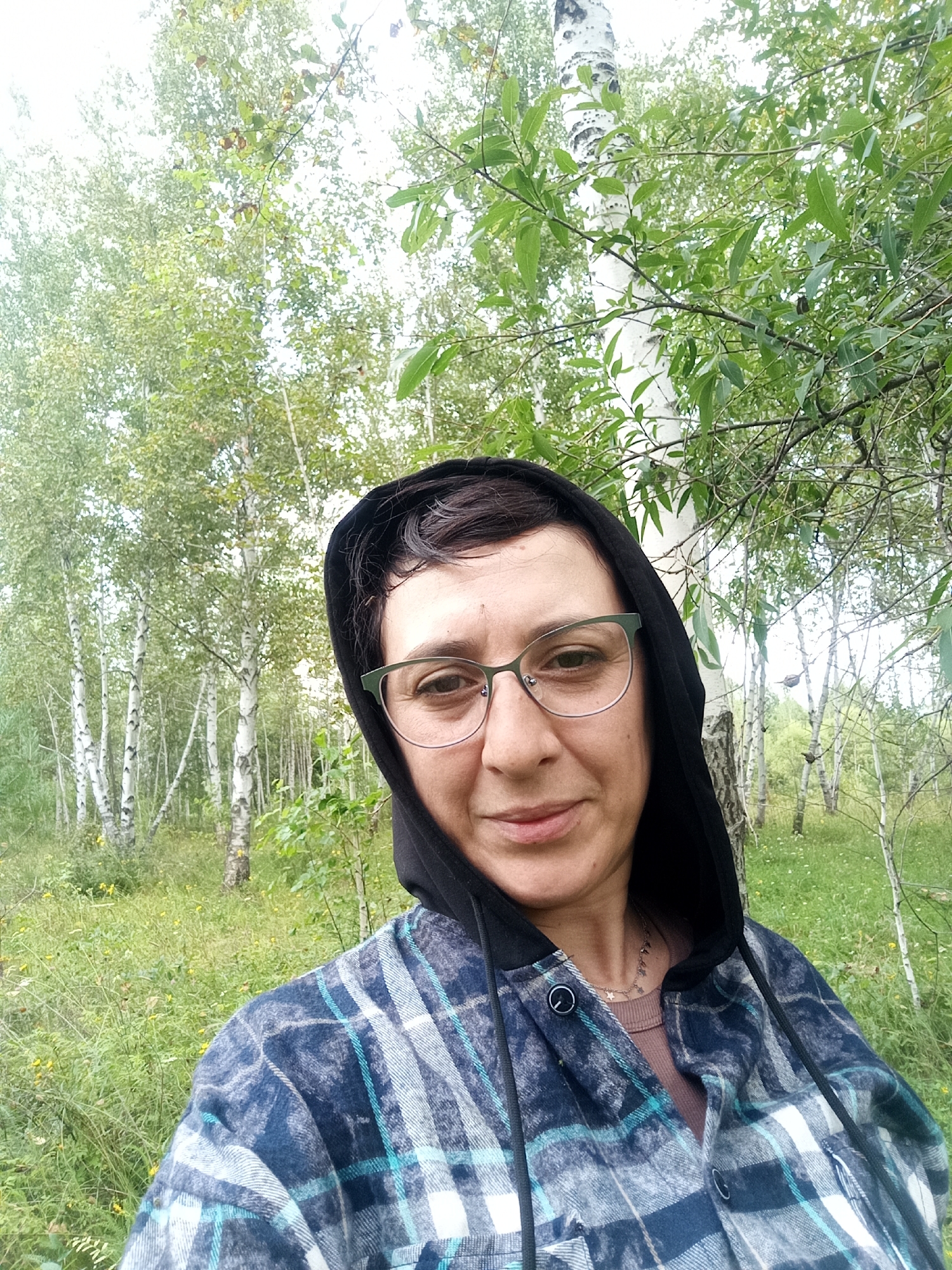 A woman wearing a black hooded sweatshirt and a blue and white plaid shirt is taking a selfie in a wooded area. She has short, dark hair and is wearing glasses. The background consists of trees and foliage, with birch trees visible. The ground is covered in grass and some small yellow flowers. The location is identified as Nikolskoye, Russia.