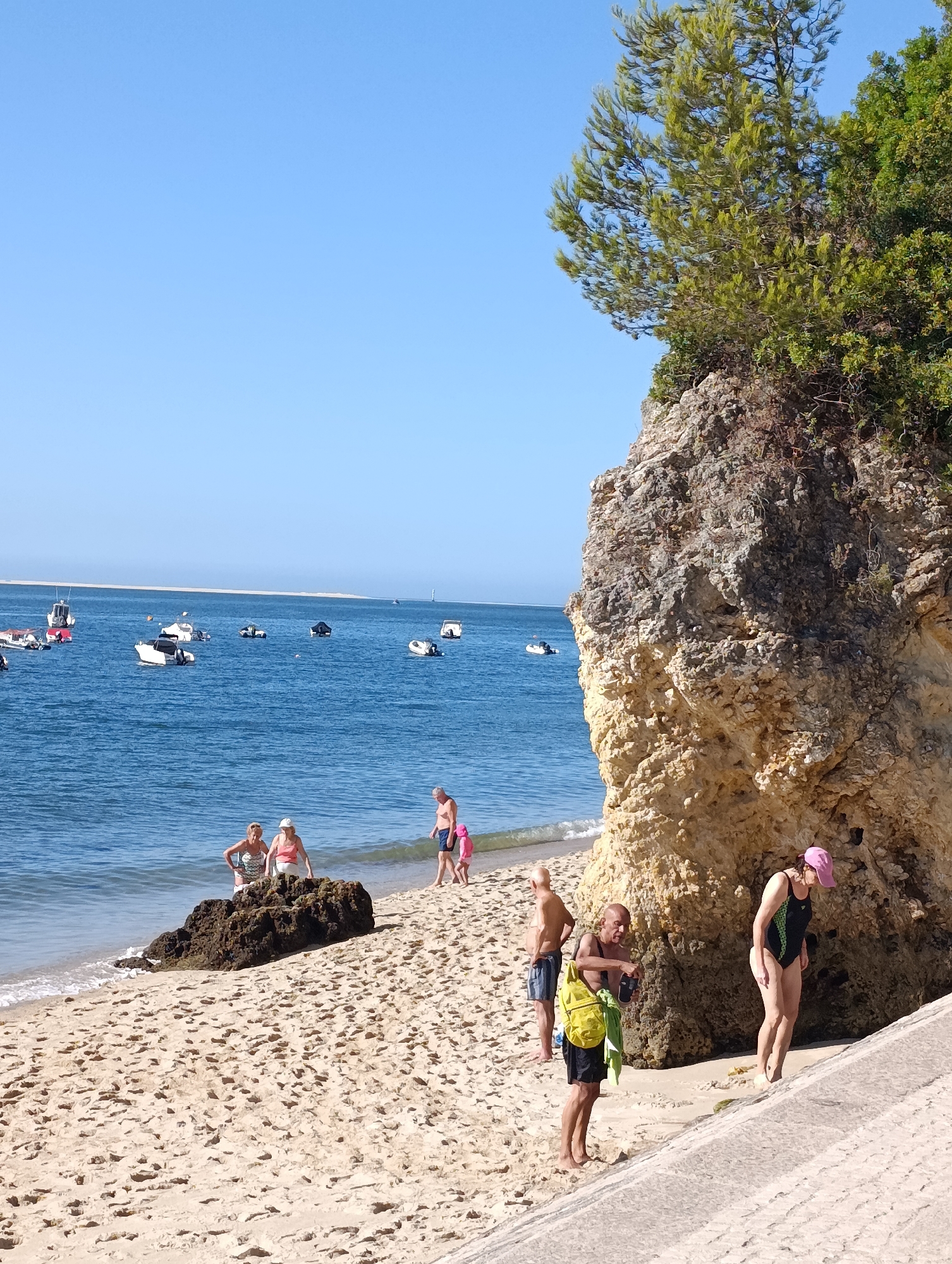 A sunny day at the beach near Setúbal, Portugal, features a rocky cliff face on the right, with vegetation growing on top. The sandy beach is populated by several individuals. In the foreground, a man wearing a black swimsuit with green accents and a pink cap walks away from the camera. Next to him, a man in black shorts and a yellow backpack is holding a pair of sandals and a phone. Further left, near a dark rock in the shallow water, two women in swimwear are standing. To the right, a man and a young child in a pink swimsuit are walking on the sand. The blue ocean stretches to the horizon, dotted with several small boats. The sky is clear and blue.