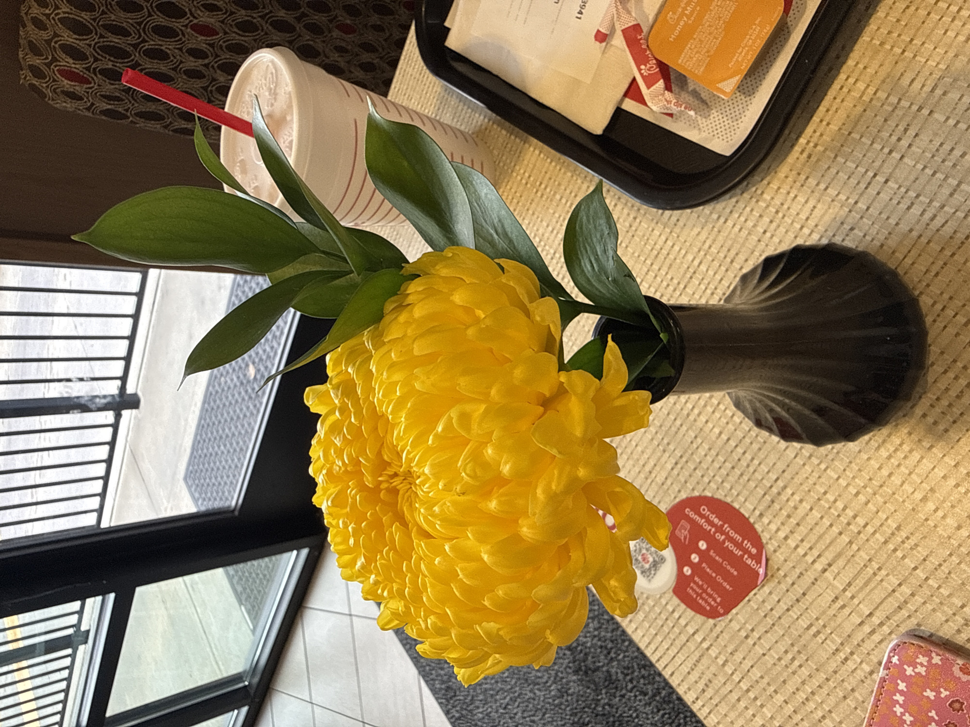 A high-angle, indoor shot features a yellow flower in a dark vase, placed on a textured beige surface. A white cup with red stripes and a red straw is visible near the flower. A black tray containing condiments such as a Chick-fil-A Honey Mustard sauce is situated next to the vase. A small, red sticker with ordering instructions is on the table next to the vase. Part of a window with a black frame is visible on the left. A corner of a wallet or small pouch is also seen in the lower right corner of the table. The location is Stone Mountain, United States of America.