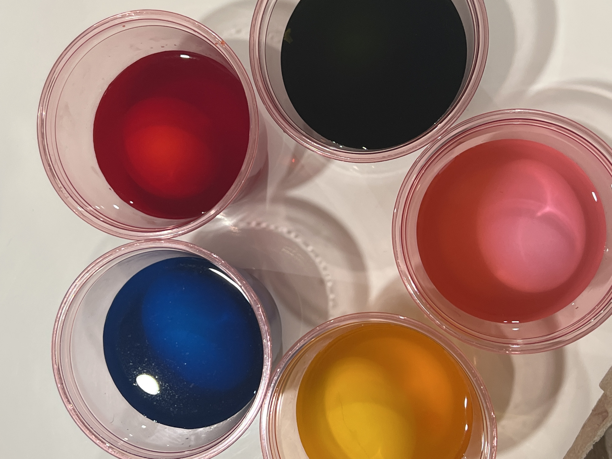 The image shows five plastic cups filled with liquid in different colors: red, black, pink, blue, and yellow. The cups are arranged in a circular pattern on a white surface. The context suggests these are dye cups for coloring Easter eggs. It is impossible to determine if this image was taken in the city of City of Newburgh, United States of America.