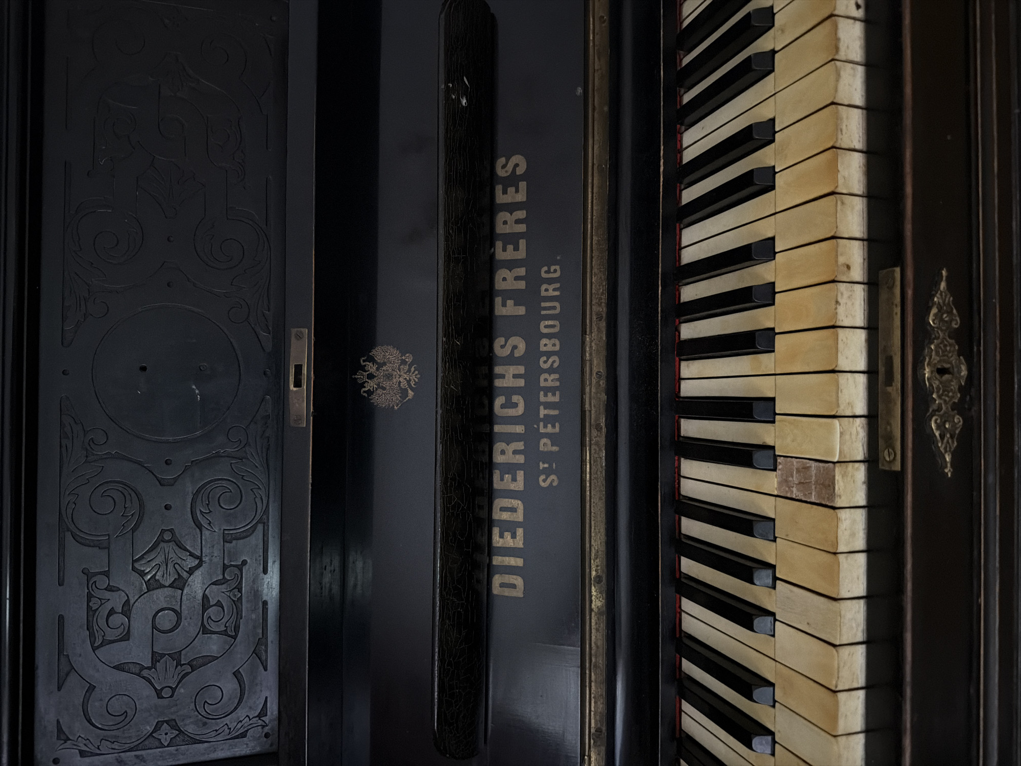 This is a close-up, eye-level, frontal view of an antique, black upright piano. The focus is on the upper portion of the piano, including the decorative carved panel at the top, the nameplate below it, and the keyboard. The carved panel features intricate, dark relief work with swirling patterns and floral motifs. Below this, centered, is a smaller, embossed emblem that appears to be a double-headed eagle, a common symbol in Russian heraldry, suggesting a connection to the Russian Empire.
The nameplate prominently displays "DIEDERICHS FRERES" in gilded lettering, with "ST PETERSBOURG." directly beneath it. This indicates the piano was manufactured by Diederichs Frères, with a likely origin or distribution point in St. Petersburg. The piano's keys are visible, with the white keys appearing aged and yellowish, and the black keys showing wear. A small, rectangular piece of material, possibly wood or ivory, is missing or discolored on one of the white keys. The overall aesthetic suggests an old, possibly historically significant instrument.
The image does not contain any people, actions, or interactions. The environment is limited to the piano itself. The location in Kolomna, Russia, is not discernible from the visual information provided in the image.