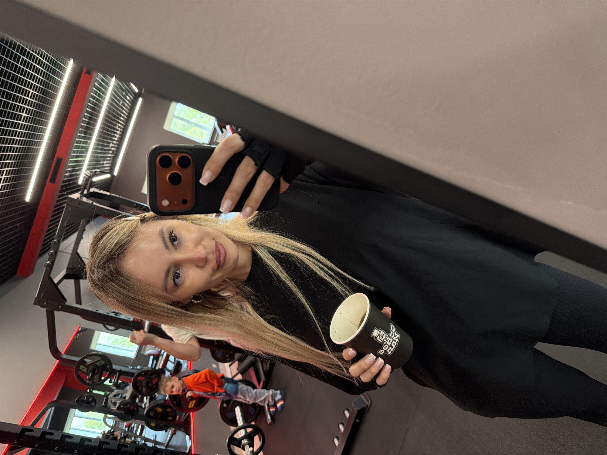A young woman with long blonde hair, wearing a black top and black fingerless gloves, is taking a mirror selfie in what appears to be a gym in Birobidzhan, Russia. She holds a black smartphone with a triple-lens camera system in her right hand, positioned to capture her reflection. In her left hand, she holds a black paper cup with visible text "COFFEE BEAT" and a graphic of a character, likely depicting a coffee-related logo. The cup appears to contain a light-colored beverage, possibly coffee, indicated by a light foam line.
The woman has a neutral to slightly smiling expression, looking directly into the phone's camera. Her nails have a light French manicure.
The background, visible in the mirror's reflection, shows a gym interior with various weightlifting equipment, including black and red power racks and barbell plates. A blurred reflection of another person, possibly a child or young person in an orange shirt, is discernible near the equipment. The gym features dark flooring, white walls, and large windows revealing green foliage outside, suggesting it is daytime. Overhead, there are grid-patterned ceiling lights. The overall setting indicates a modern fitness facility.