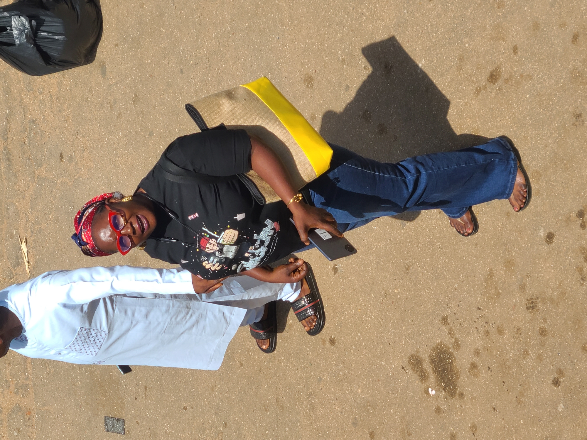 A high-angle shot captures two individuals walking on an asphalt surface, likely in Jos, Nigeria. On the left, a person wearing a white long-sleeved tunic with intricate patterned pockets and dark slide sandals walks away from the camera. To the right, a woman with a red headscarf, large red-rimmed sunglasses, and a black t-shirt with a graphic print is walking towards the camera. She wears blue jeans and dark slide sandals, and carries a large beige and yellow tote bag. Her right arm is adorned with a gold bracelet. In the background, a large black garbage bag is visible. The ground shows signs of wear and tear, with scattered oil stains and debris. The lighting suggests it is daytime, with the sun casting distinct shadows.