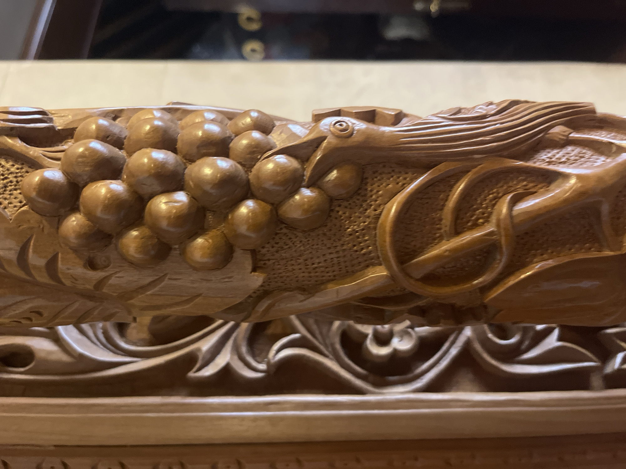 The image shows a close-up of a carved wooden column. The carving features a cluster of grapes, a bird's head with an eye, and what appears to be a circular object with a cross shape within it, possibly an anchor or a stylized wheel. The wood is polished and has a warm, reddish-brown hue. The background is out of focus but suggests an indoor setting with light-colored walls and darker elements, possibly furniture or doorways. The detail of the carving suggests it is part of a decorative piece, such as a furniture element, a pillar, or an architectural feature. The location in Foini, Cyprus, is noted for its tradition of wood carving, making this a plausible piece from the region.