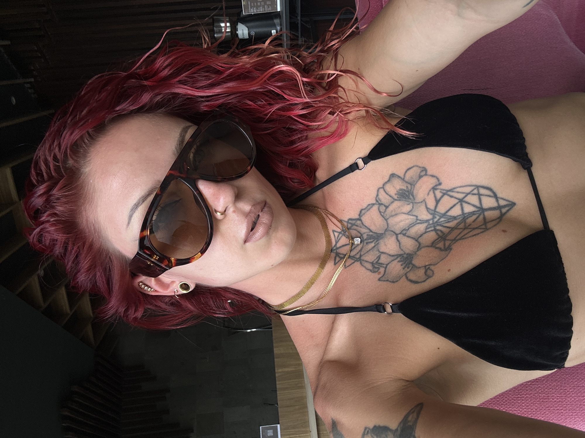 A fair-skinned person with red, curly hair lies on a pink surface, wearing a black bikini top. The person has multiple ear piercings and is wearing brown sunglasses. A floral tattoo is visible on the upper chest. The person is also wearing a gold necklace. A partial view of a tattooed arm is visible in the lower right corner of the image. The background is blurred and indistinct, with glimpses of dark surfaces and what appears to be a window reflecting light. The location is unspecified in the prompt.