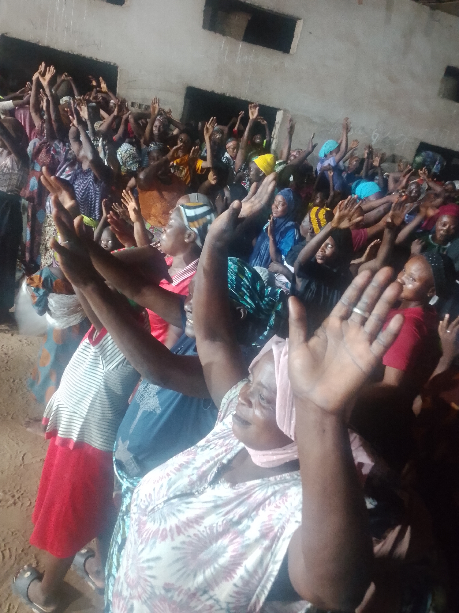 In an indoor environment with a dirt floor, a crowd of people, predominantly women, are gathered. Many of them have their arms raised, some with hands open, in what appears to be a gesture of worship or prayer. The individuals are wearing various types of clothing, including patterned fabrics and headscarves. The lighting suggests it is either evening or an indoor space with limited illumination. The background shows a rough, unfinished wall. The scene captures a moment of collective spiritual or emotional expression.