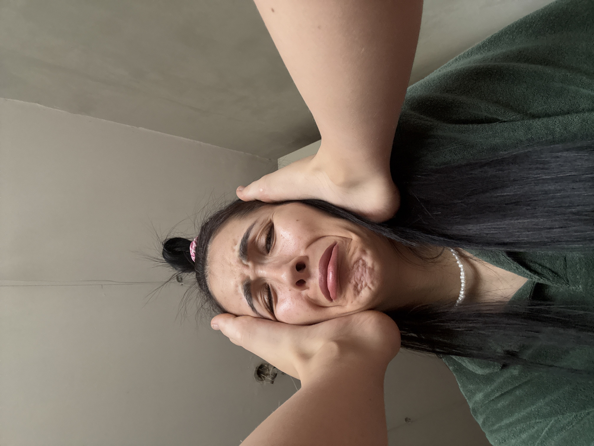 An indoor, low-angle portrait captures a woman's face being playfully pressed by two bare feet, against a backdrop of plain, light-colored walls. The woman, with long dark hair styled in a small bun secured by a pink tie, wears a dark green textured garment, possibly a robe or towel, and a pearl necklace. Her face is contorted in an exaggerated expression of distress or discomfort, with furrowed brows, squinted eyes, and her mouth pulled down to one side, suggesting a theatrical or humorous reaction rather than actual pain. One bare foot rests on her forehead, while the other presses firmly against her chin and jawline, distorting her features. The lighting is even, suggesting an interior space without strong natural light sources visible. No text is visible in the image. This scene takes place in Votkinsk, Russia.