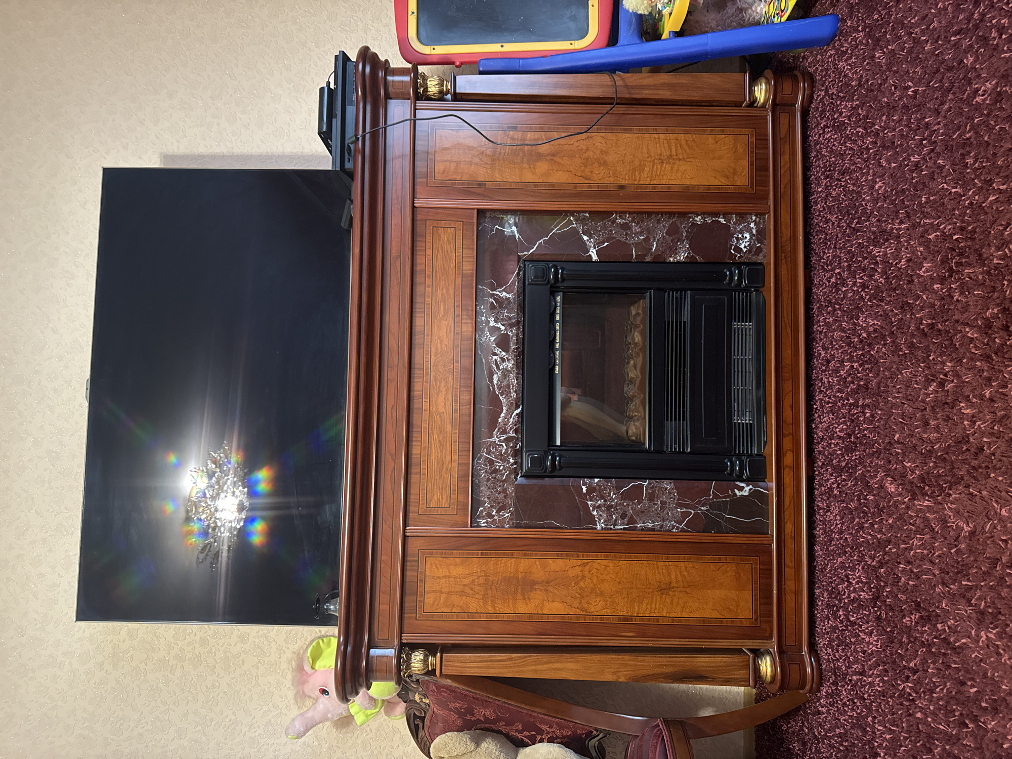 A television is perched atop a wooden fireplace mantel. The mantel is ornate, with dark wood trim and lighter wood panels. The fireplace insert itself is black, with a grate at the bottom. A chandelier is reflected in the television screen, casting rainbow-colored light. To the left of the fireplace, a plush pink elephant toy is visible, and part of a chair with a burgundy patterned fabric. To the right, a blue and yellow easel-like object is standing. The fireplace is situated on a dark red, shag carpet. The wall behind the television has a textured, light-colored wallpaper with a subtle floral pattern. A black electronic device, possibly a game console, is placed on top of the mantel to the right of the television, connected to a power cord.