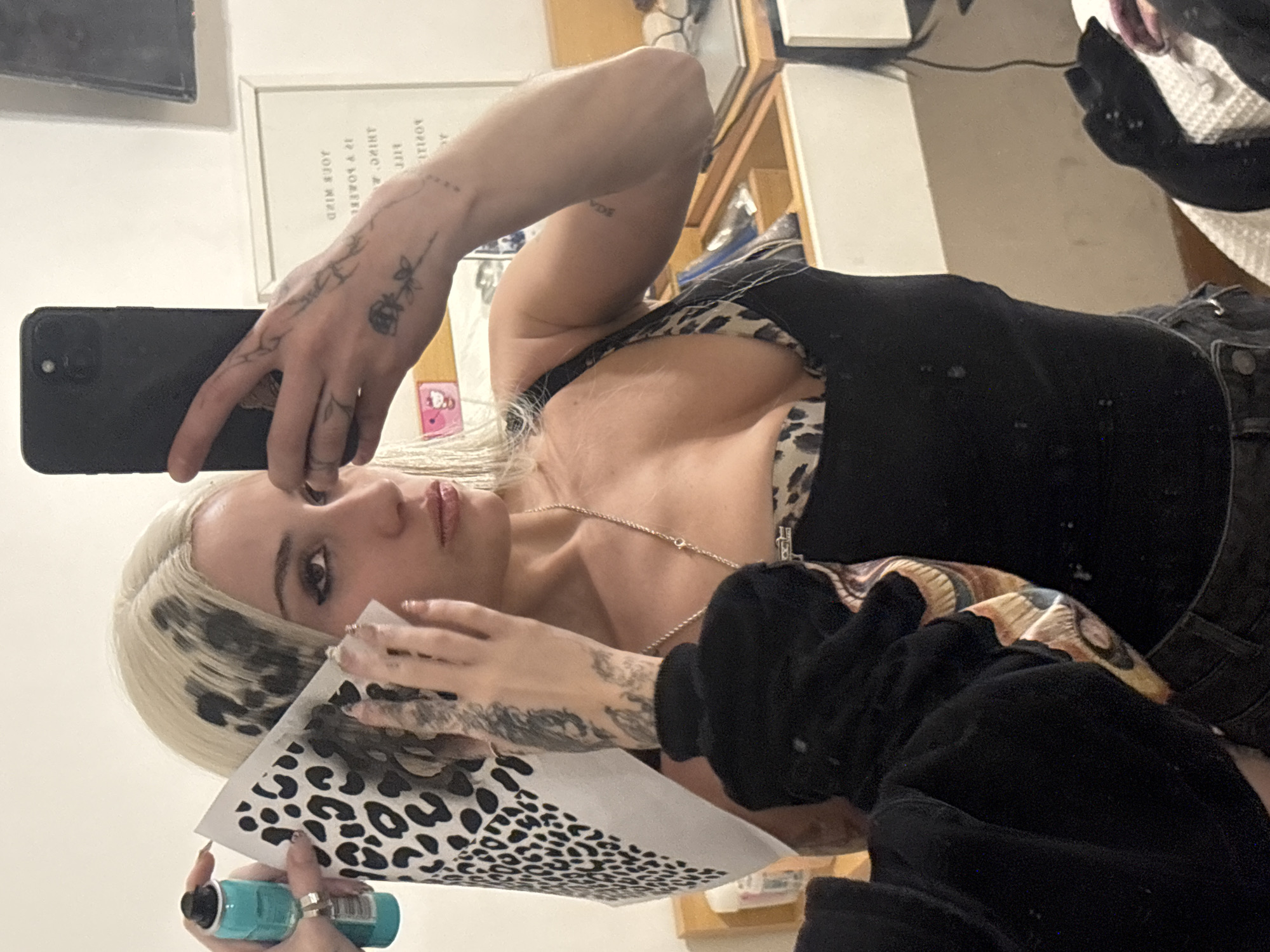 The image captures a blonde woman taking a selfie in a mirror, holding a black smartphone with her right hand. She is engaged in a creative activity, holding a piece of paper with a black leopard print pattern and a small teal/green spray bottle in her left hand, suggesting she might be applying the print to her hair, which already shows distinct black leopard spots on her blonde locks.
The woman is wearing a black tank top over a leopard print bra or bralette, and black jeans. A black long-sleeved garment covers her right forearm. She has numerous fine-line tattoos on her hands and arms, including floral/vine patterns, a small rose on her left hand, and a word that appears to be "WIDE" on her upper right arm. A delicate gold chain necklace is visible around her neck, and her nails are long and manicured.
The setting is an indoor space, likely a room with white walls and wooden shelves or furniture in the background. Reflected in the mirror, behind her head, is a white-framed picture containing reversed text. Visible words include "POSITIVITY" and parts of what appears to be an inspirational quote, with "YOUR MIND" and "IS A POWERFUL" being somewhat legible. There are no specific cues for time of day or weather; the lighting suggests a well-lit interior. The woman's expression is neutral and focused. The image is contextualized as being taken in Buenos Aires, Argentina.