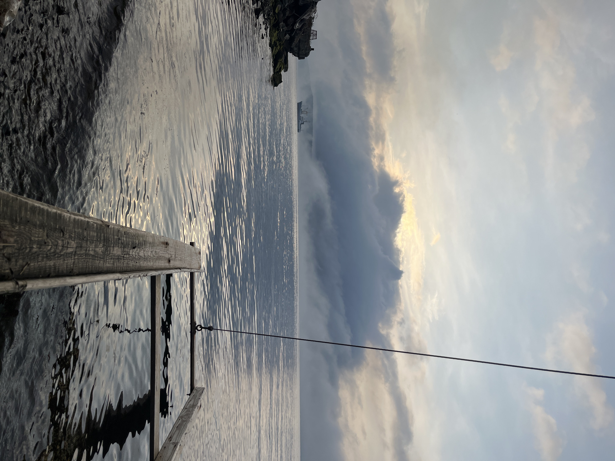 The image depicts a serene waterscape at dawn, likely in Sovetskaya Gavan', Russia, given the context of the city. The sky is filled with a mixture of soft clouds, with hints of golden light breaking through, suggesting the early morning sun. A large cargo ship is visible in the distance, partially obscured by a dense layer of fog or low clouds that hug the horizon and the distant shoreline.
In the foreground, the calm surface of the water reflects the muted colors of the sky. To the left, a rocky embankment with some debris, including what appears to be old tires and metal structures, marks the edge of the land. The water laps gently against this shore.
Dominating the right side of the frame is a wooden structure partially submerged in the water. This structure appears to be a makeshift dock or platform, constructed from weathered wooden planks. A dark rope hangs from above, attached to a metal ring on this structure, suggesting it might be a mooring point or part of a larger system. The water beneath this structure is dark and reflects the wooden beams above, hinting at its depth. The overall atmosphere is tranquil and slightly misty, characteristic of a coastal morning in a port city. There are no people visible in the image.