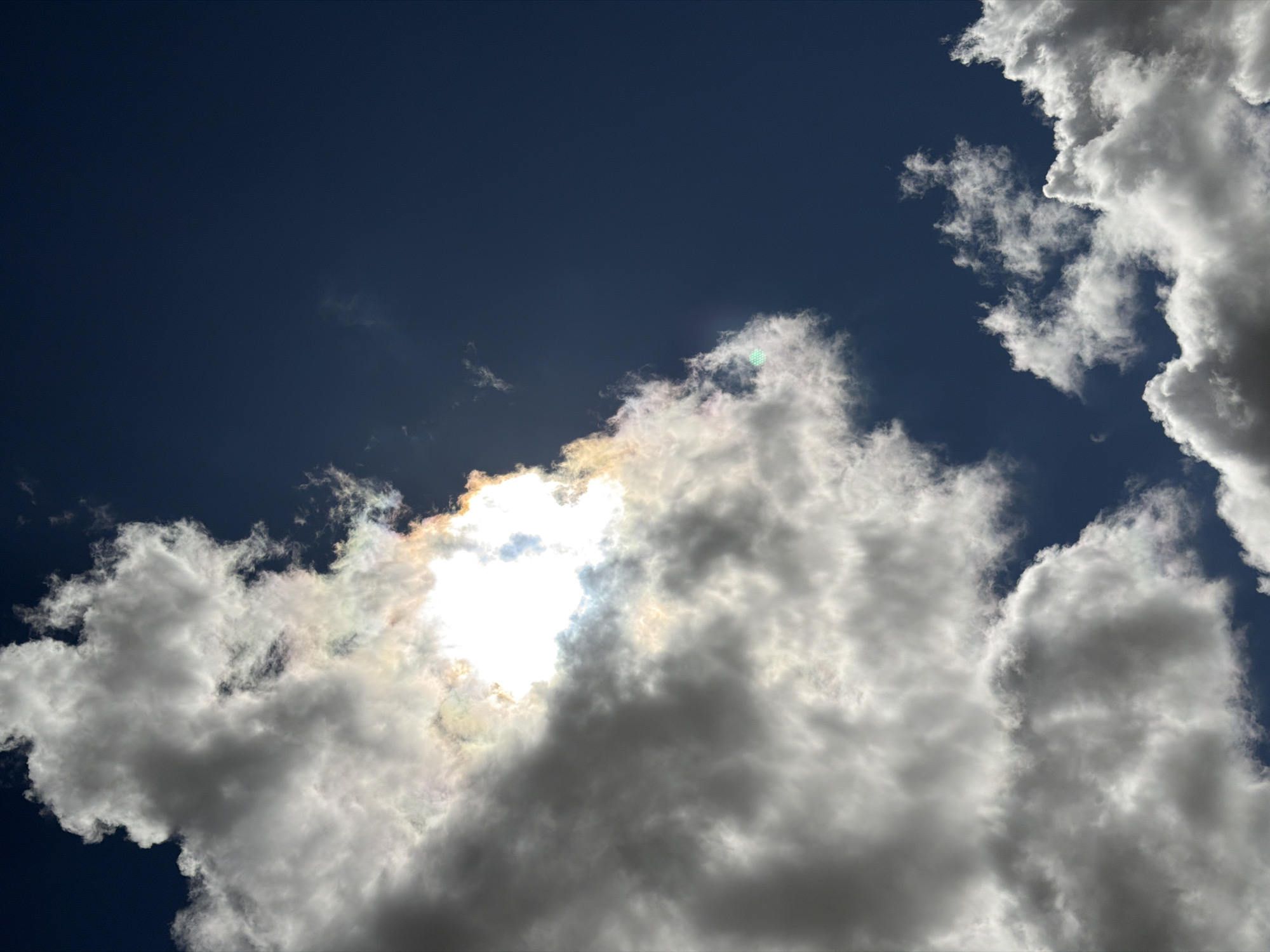 The image shows a view of clouds in the sky. The sky is a deep blue color in the upper left portion of the frame. The clouds are white and billowy, and are concentrated in the lower half and the right side of the image. The sun is behind the clouds, creating a bright spot and illuminating the edges of the clouds. There are hints of rainbow iridescence near the sun. The image was taken during daylight hours. There are no people, buildings, or specific environmental features visible that would definitively locate the image in Rossiyskiy, Russia.