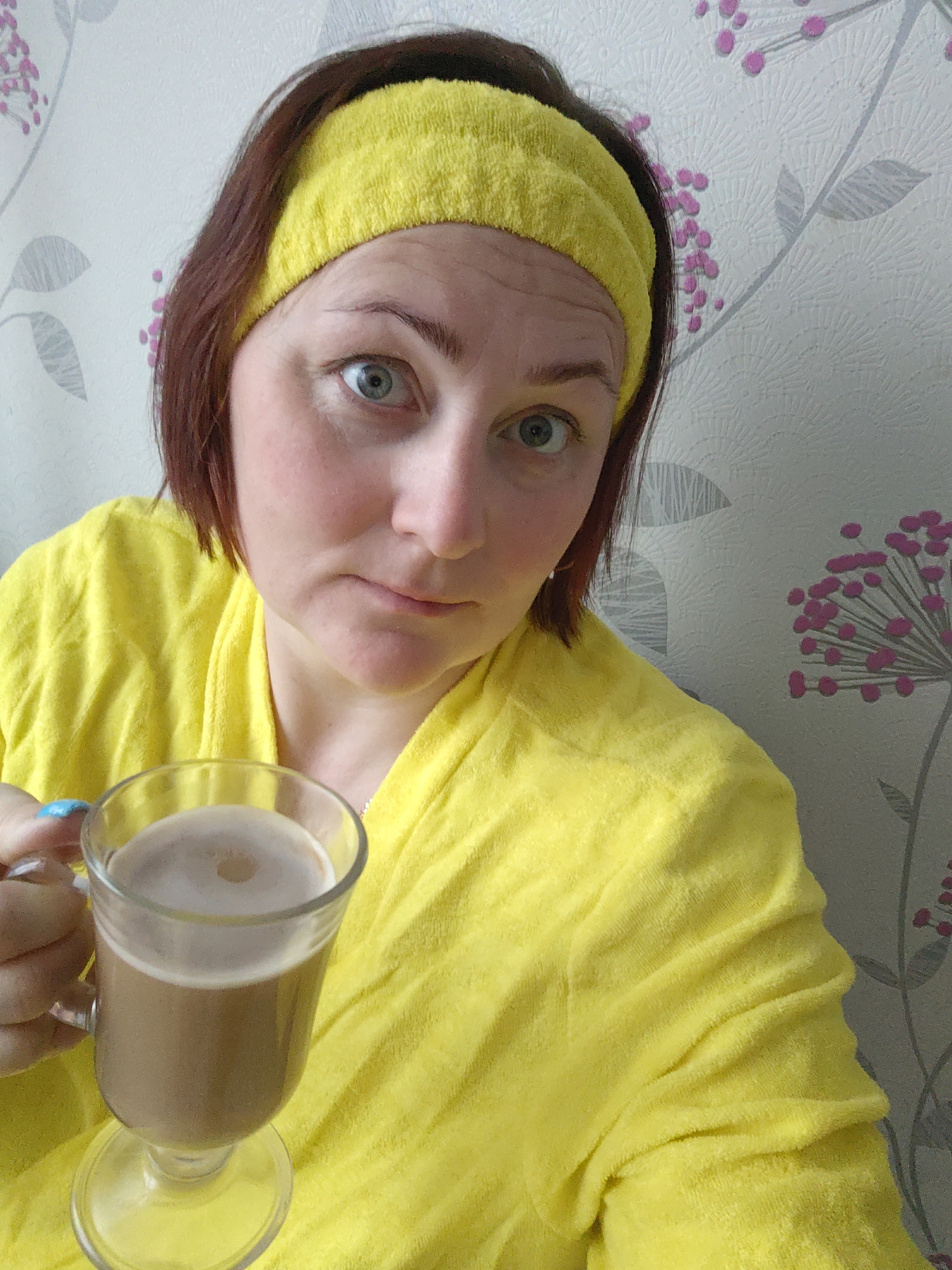 A woman with fair skin, shoulder-length auburn hair, and a yellow terrycloth headband and bathrobe is holding a glass of coffee. She has blue eyes and is looking directly at the camera with a slightly quizzical expression. The background is a wall with a floral pattern featuring gray leaves and pink flowers. The location is not discernable from the image.