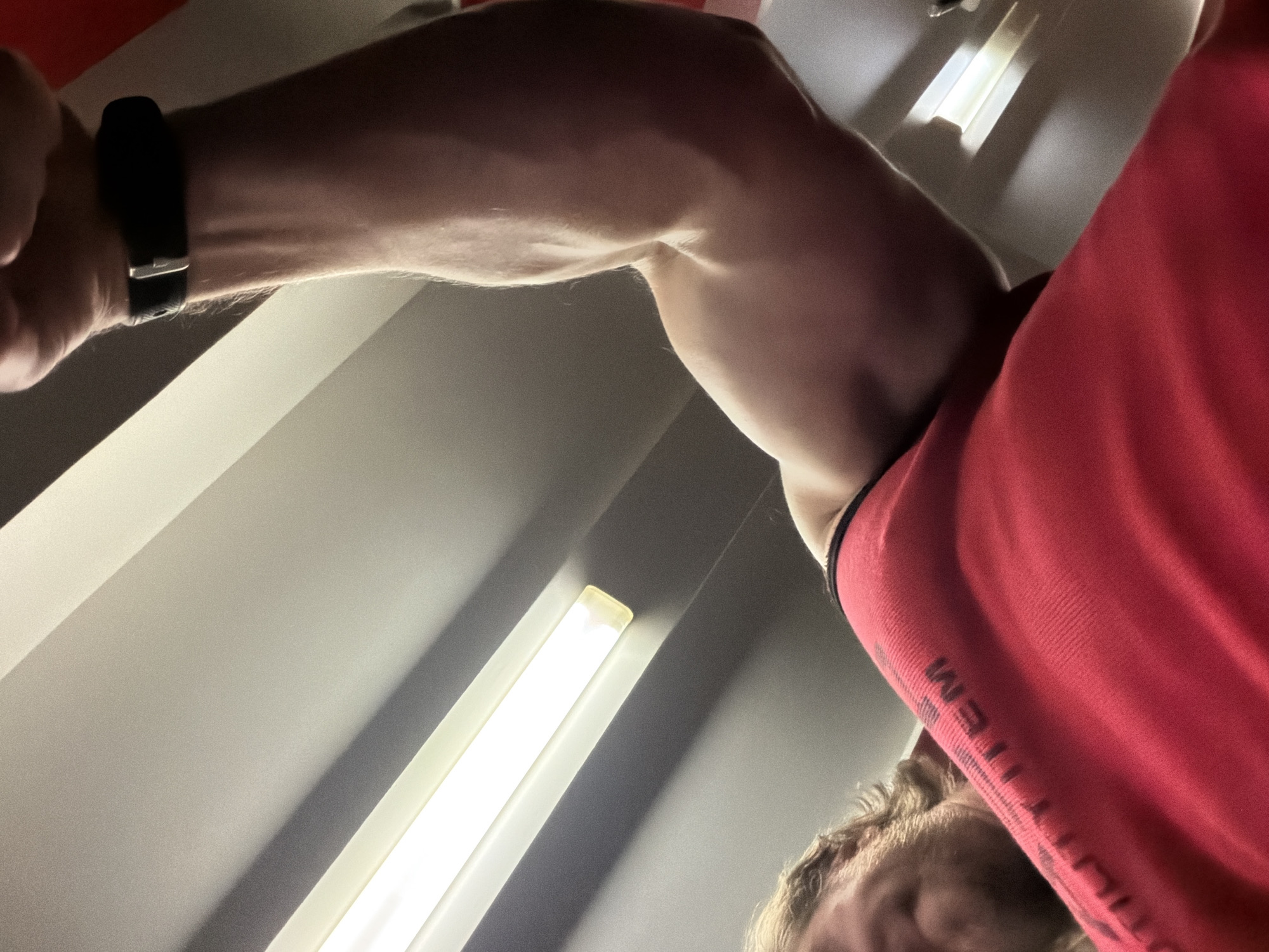This image captures a low-angle perspective of an individual flexing their right bicep within an indoor setting in Saint Petersburg, Russia. The subject is a fair-skinned male, partially visible in the upper left, featuring light brown hair and facial stubble or a short beard. He is wearing a red sleeveless top or tank top with dark lettering on the left shoulder, partially readable as "ULITEM," likely part of a larger word such as "PROUTILITEM." His right arm is the primary focus, extended across the frame from the lower left to the middle right, showcasing muscle definition and vascularity in the bicep and forearm. A black wristband or smartwatch with a silver buckle is worn on his right wrist. The background consists of a plain, light-colored (off-white or light gray) ceiling and upper wall. Several fluorescent tube light fixtures with diffusers are visible diagonally across the ceiling, emitting bright white light and casting shadows. The precise location within Saint Petersburg, such as an exact address or specific type of establishment (e.g., gym), cannot be determined from the generic interior architecture and lighting visible in the scene.