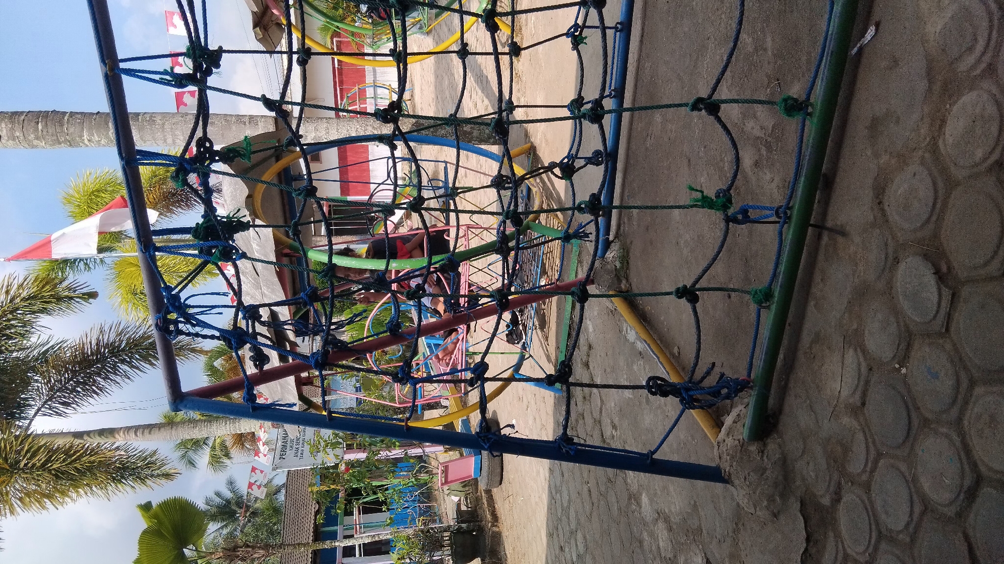An outdoor, low-angle shot captures a playground in the city of Bangun Rejo, Indonesia. The foreground is dominated by a blue, red, and green net climber, constructed of thick ropes knotted together. Beyond the net, a pink and yellow slide is visible, along with other playground equipment like swings. A white and red Indonesian flag flies from a pole in the background, framed by palm trees with lush green fronds. The ground is a mix of dirt and hexagonal paving stones. In the middle ground, two individuals are seated on a swing. The scene is illuminated by bright daylight, suggesting it is daytime.