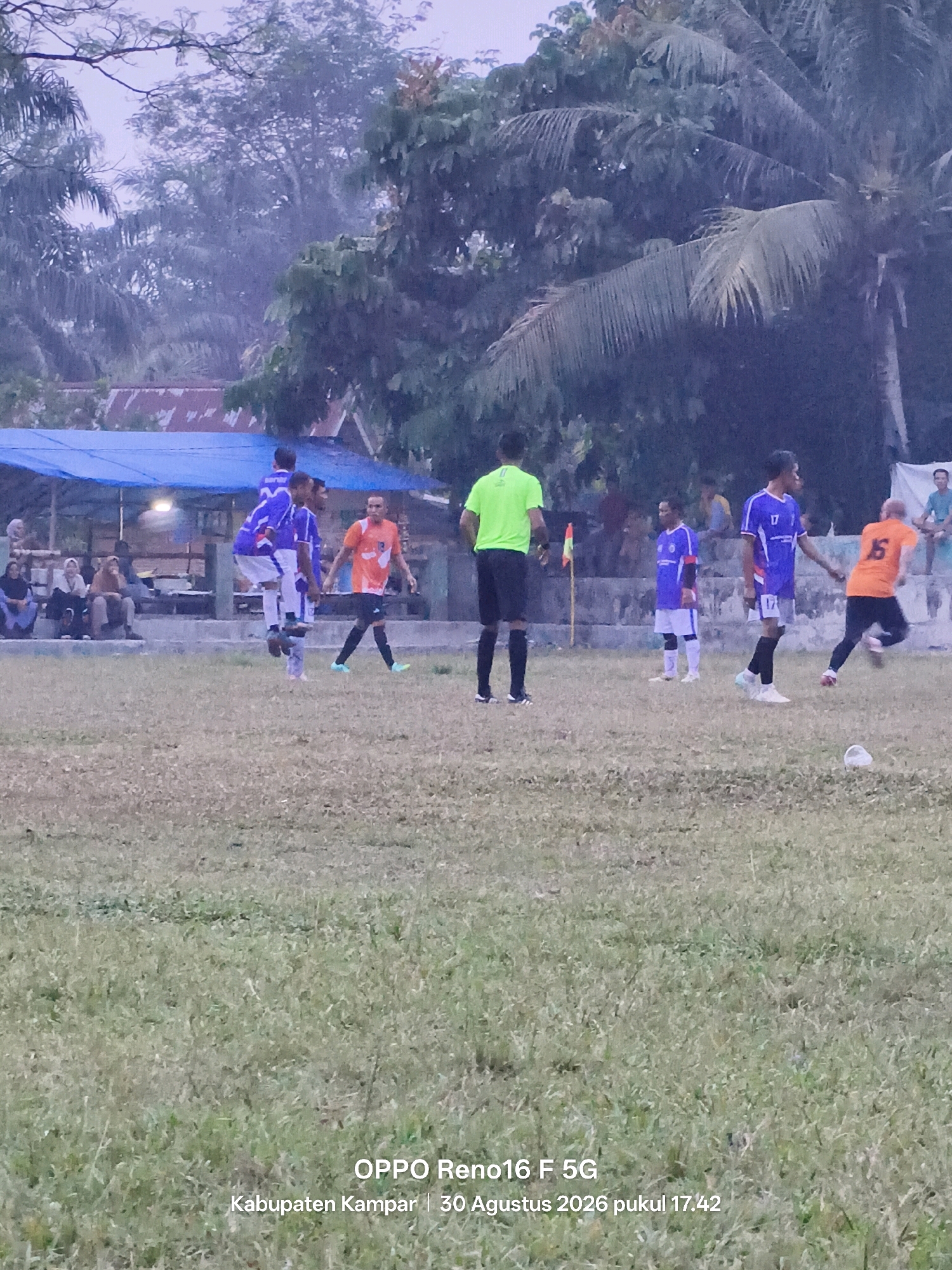 The image depicts an outdoor soccer match at a field in Simalinyang, Indonesia, on August 30, 2026, at 17:42. The field is covered in dry grass, and the sky appears overcast. Several players are visible in their team uniforms. One player in an orange jersey with the number 16 on his back is running towards the right of the frame. Another player in a blue and white jersey with the number 17 is facing towards the right and appears to be interacting with the player in orange. There are other players in blue and white uniforms scattered on the field, some in action and others standing. In the background, there are spectators sitting on a raised area, some under a blue tarp. Trees and palm trees are present in the background, suggesting a tropical or subtropical environment. The overall atmosphere suggests a casual sporting event.