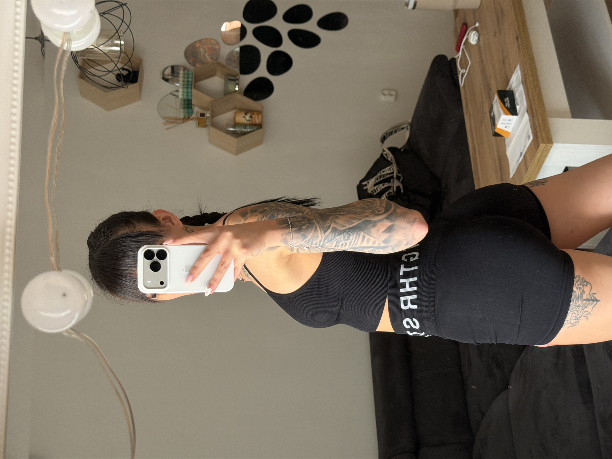 A young woman with dark hair in a braid, wearing a black athletic crop top and black shorts, is taking a mirror selfie. She has numerous tattoos on her arm. She is standing in what appears to be a living room or bedroom, with a dark sofa and a wooden coffee table visible. The room has white walls, with decorative items on shelves and a wall decal. The image is captured in a mirror reflection, and no specific location in Plovdiv, Bulgaria can be identified.