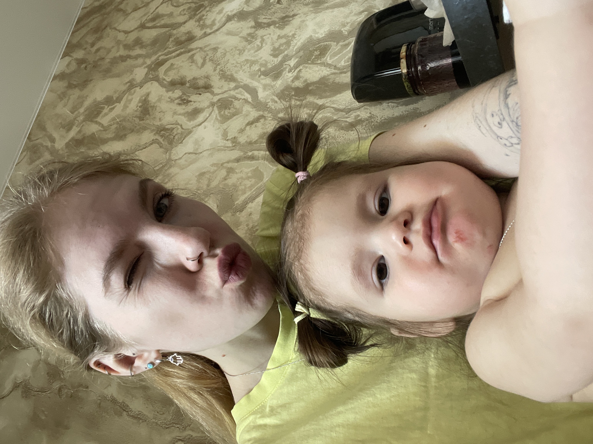 A selfie captures a young woman and a toddler. The woman, with blonde hair, winks with one eye closed and puckers her lips. She has multiple earrings and a nose ring. The toddler beside her has brown hair in pigtails tied with pink and yellow ribbons, and a neutral expression. They are both wearing light green shirts. The woman has a tattoo on her arm. In the background, there's a beige patterned surface and a black object with a glass jar, possibly containing liquid. The scene is indoors. The location is not determinable from the image.
