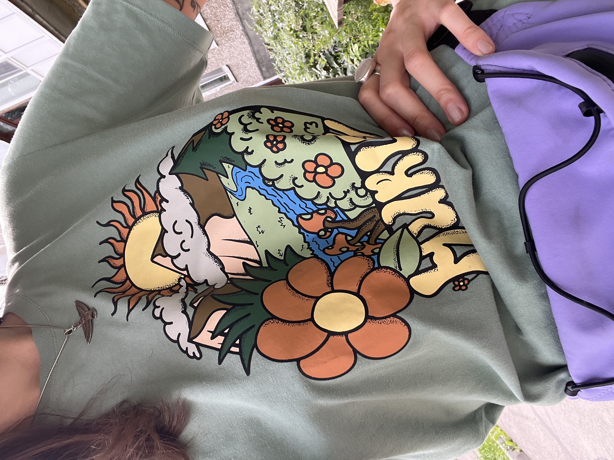 A close-up, medium shot shows a person wearing a light green t-shirt with a colorful graphic on it and a purple fanny pack around their waist. The t-shirt features a stylized depiction of a landscape with a sun, clouds, mountains, a river, trees, mushrooms, and flowers, along with the word "HIKR" in yellow lettering. A silver bird pendant is visible on a chain around the person's neck, and a silver ring is on their right index finger. The background is out of focus but appears to be an outdoor setting with green foliage and a building. The image is set in Gubakha, Russia.